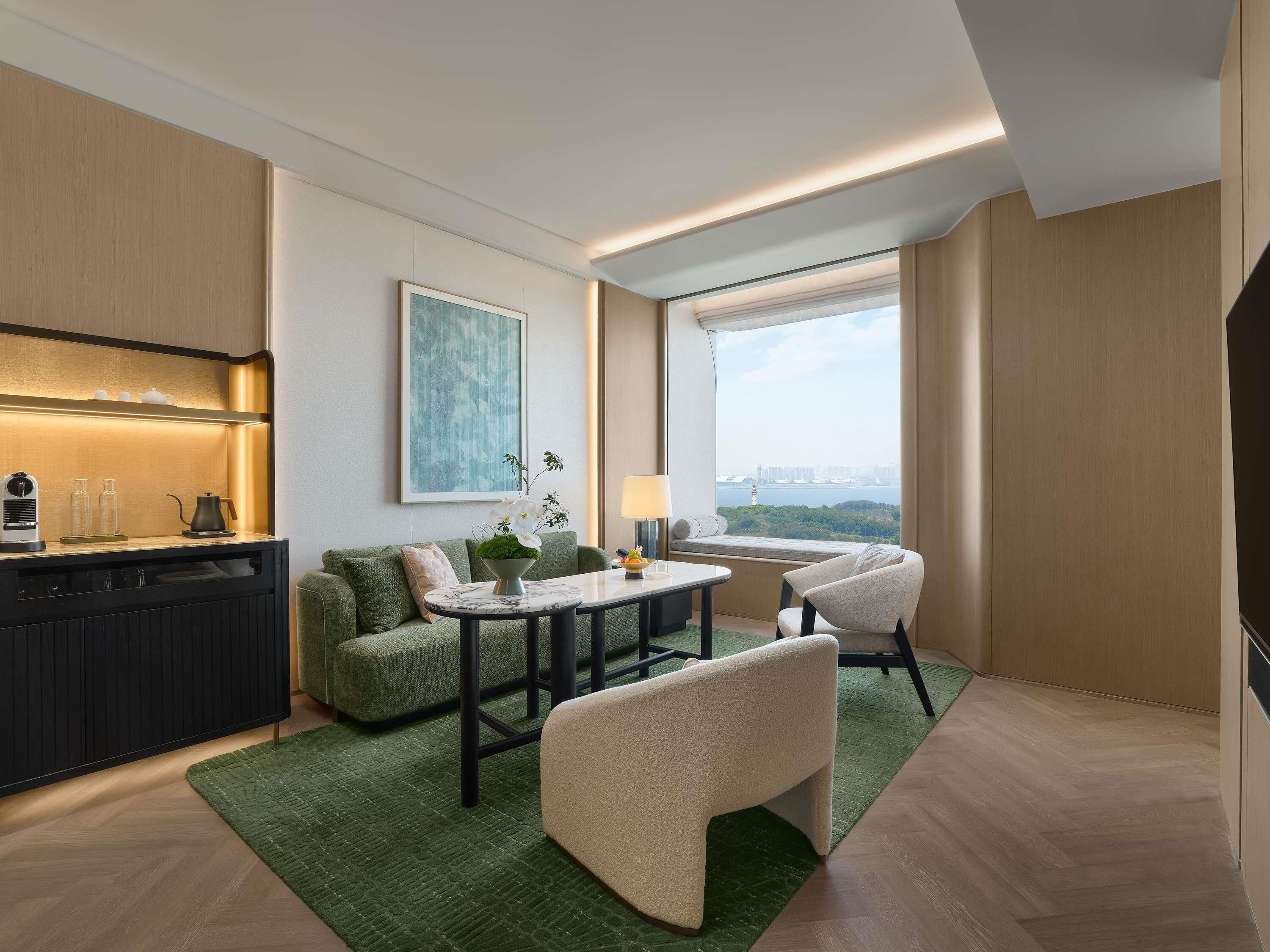 A modern hotel room with a kitchenette, a dining area, a sofa, a chair, and a large window offering a city view.