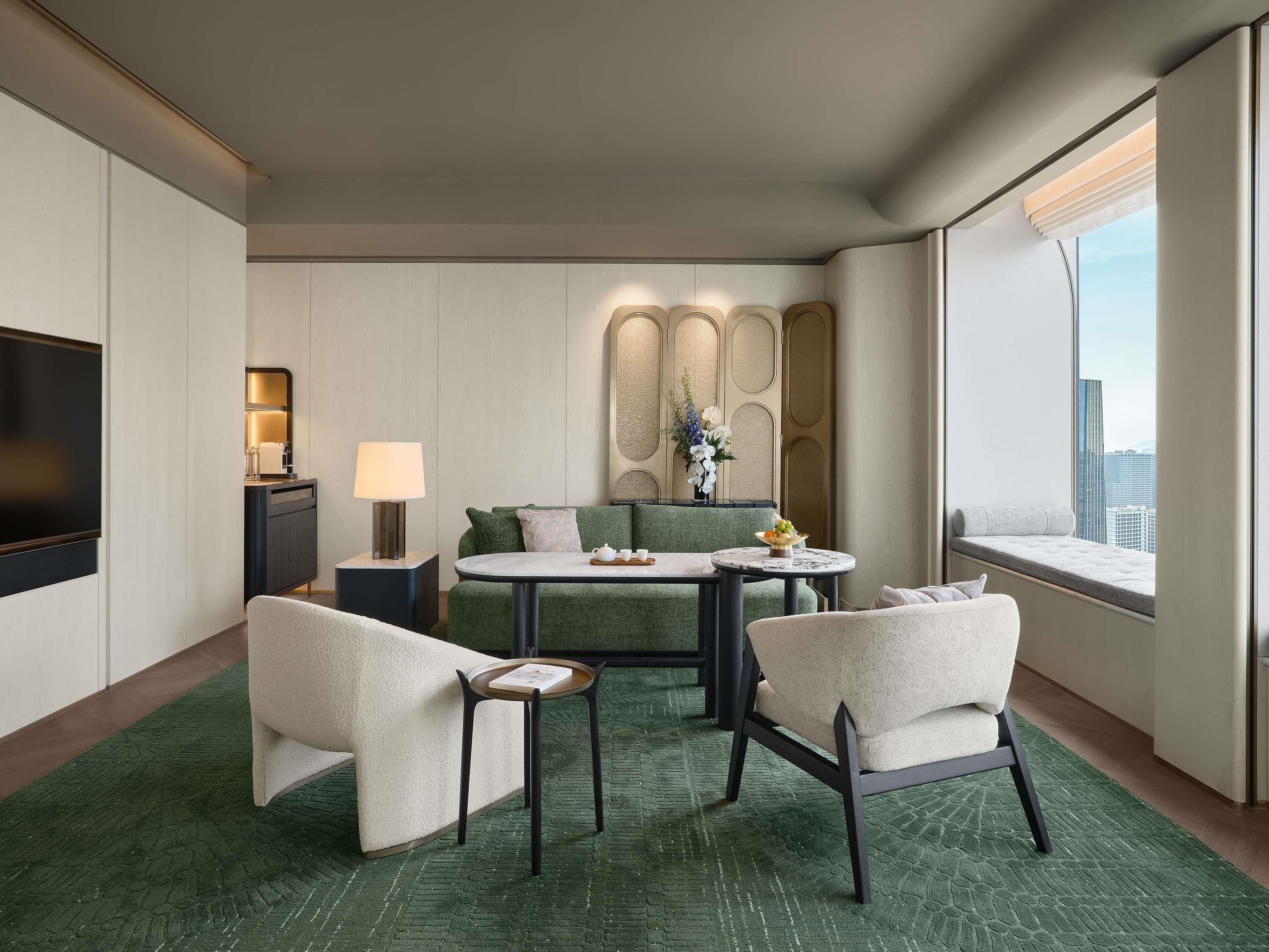 Habitación de hotel con televisor de pantalla plana, un sofá, una mesa de comedor con sillas, una lámpara y un panel decorativo en la pared.