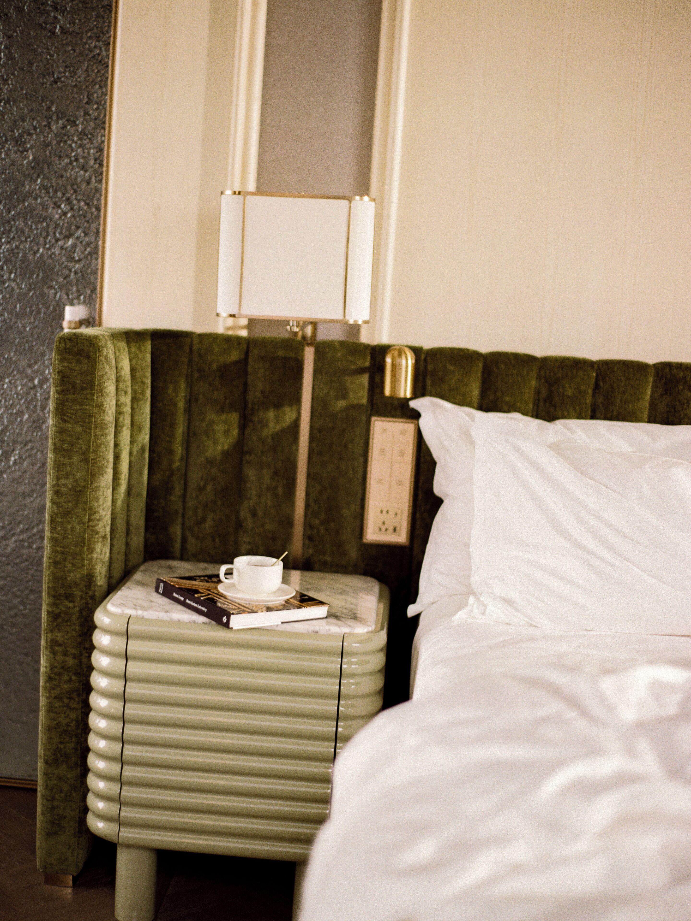 Habitación de hotel con una cama, un cabecero tapizado en verde, una mesita de noche con una taza, un libro y una lámpara.