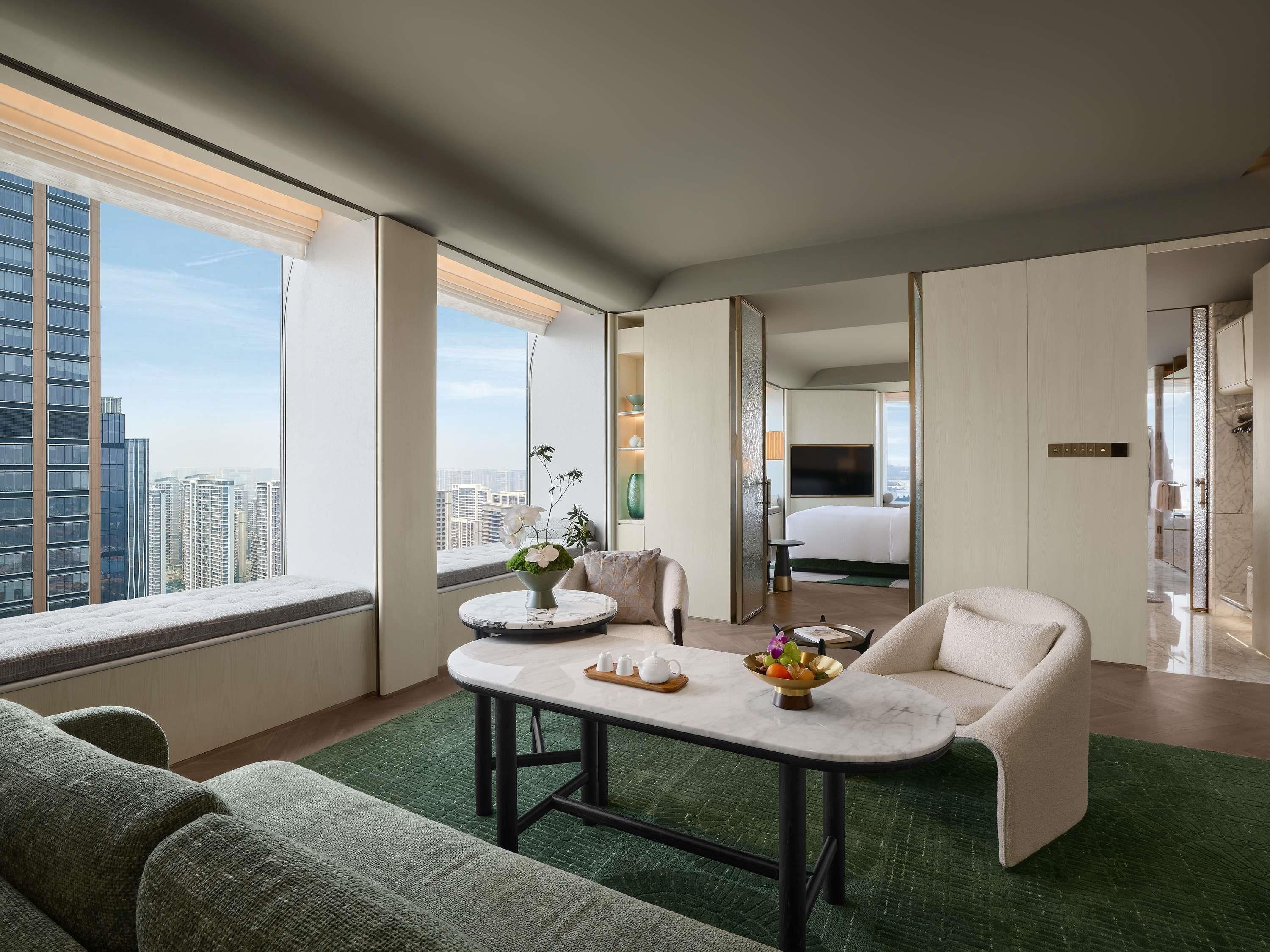 Een moderne hotelkamer met uitzicht over de stad, een zithoek met een tafel, en een afzonderlijke slaapkamer met een bed en een televisie.
