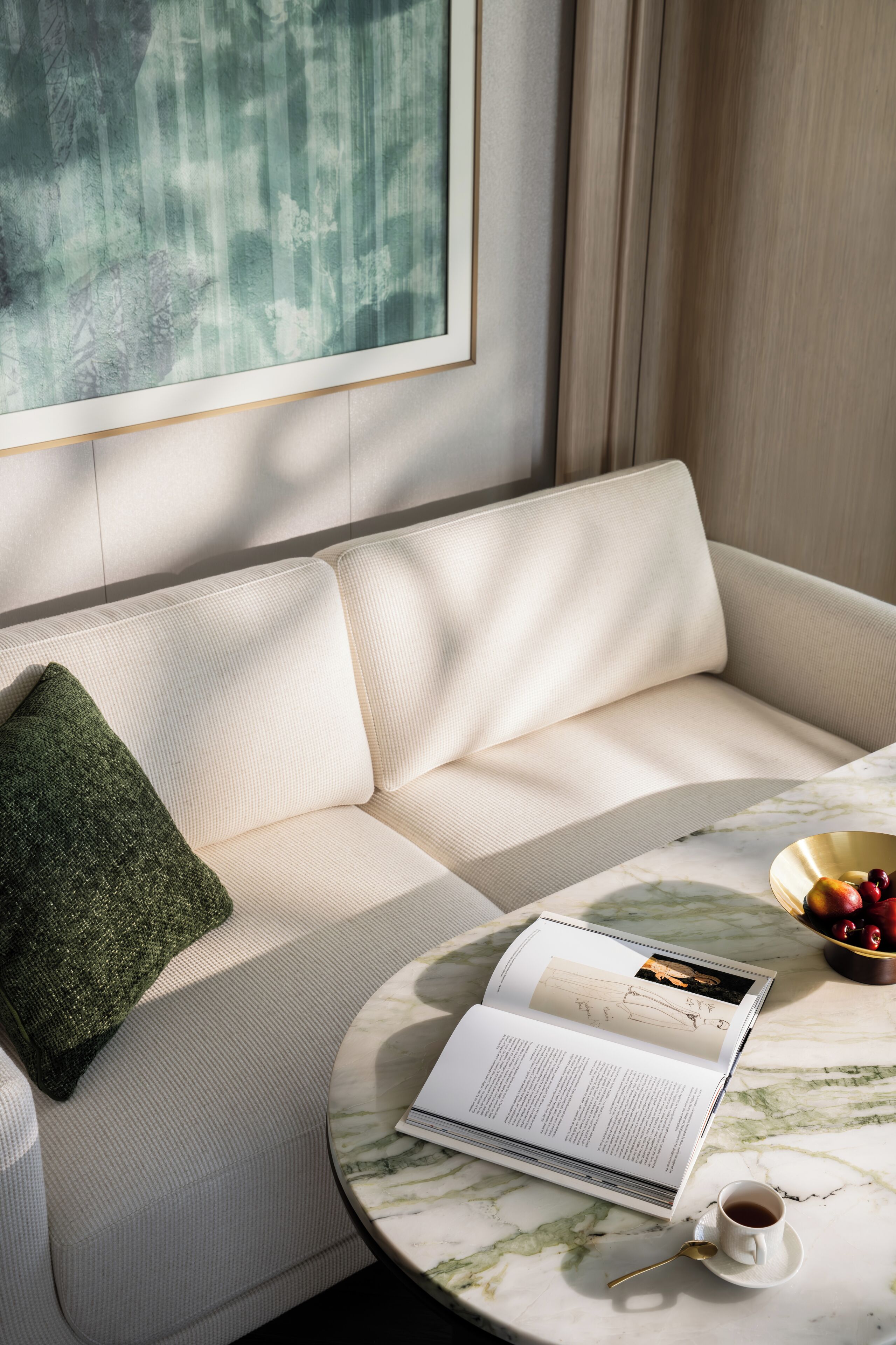 Un salón moderno con un sofá color beige, un cojín verde, una mesa de centro de mármol, un libro, una taza de café y un frutero.