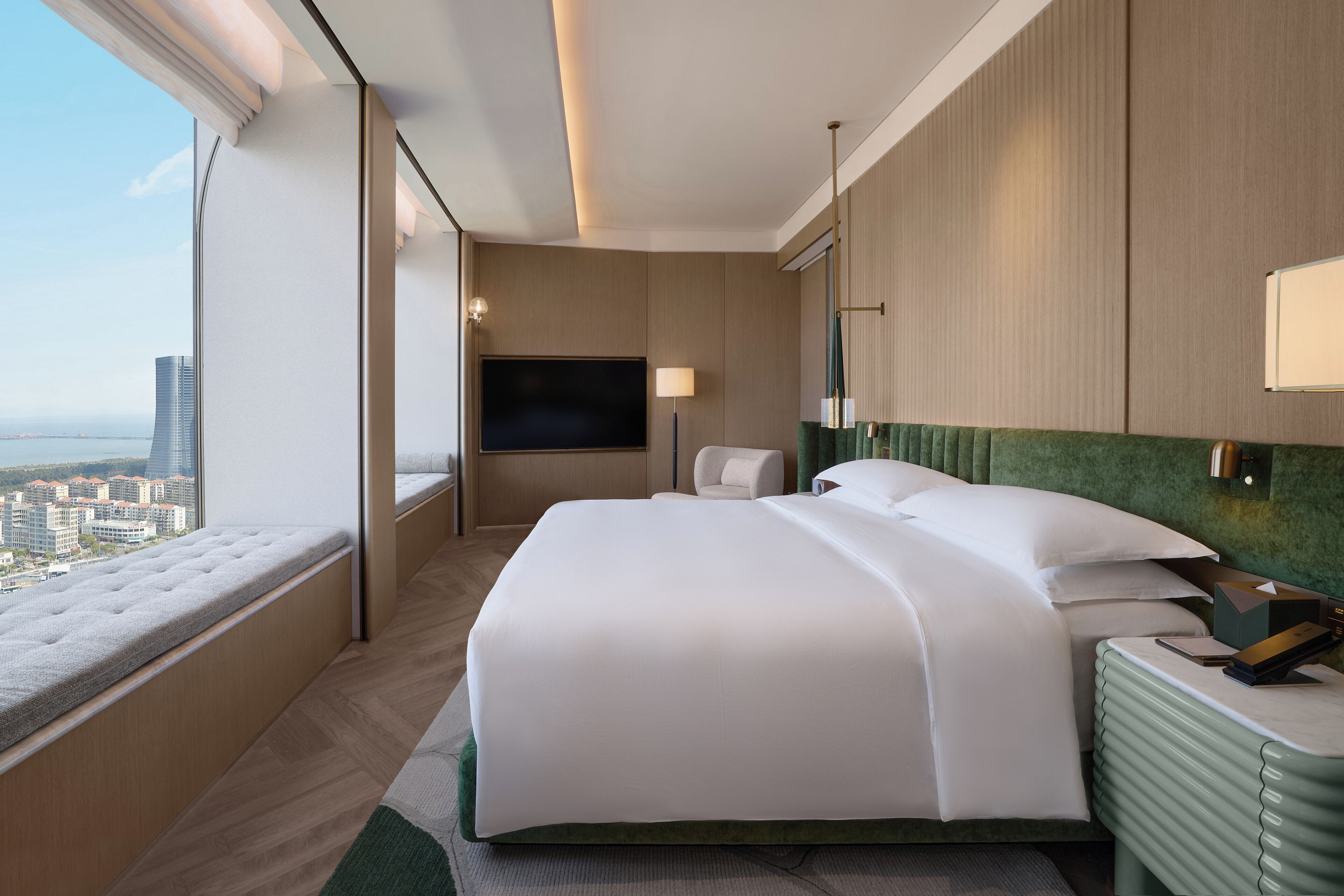 A hotel room with a large bed, a TV, a green headboard, and a view of the city.