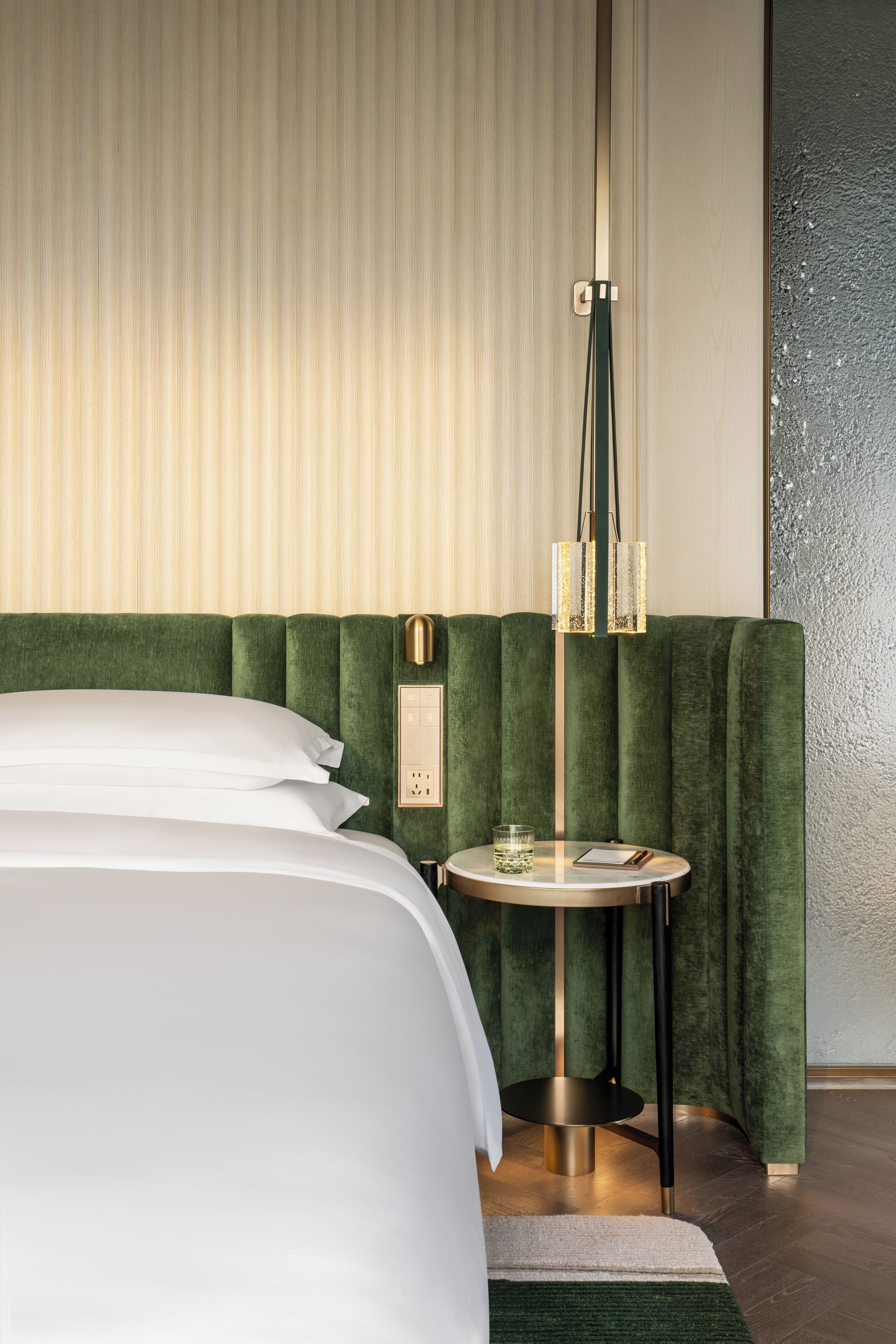 Une chambre d’hôtel moderne avec un grand lit, une table d’appoint ronde, une tête de lit verte recouverte de tissu et un mur à motifs.