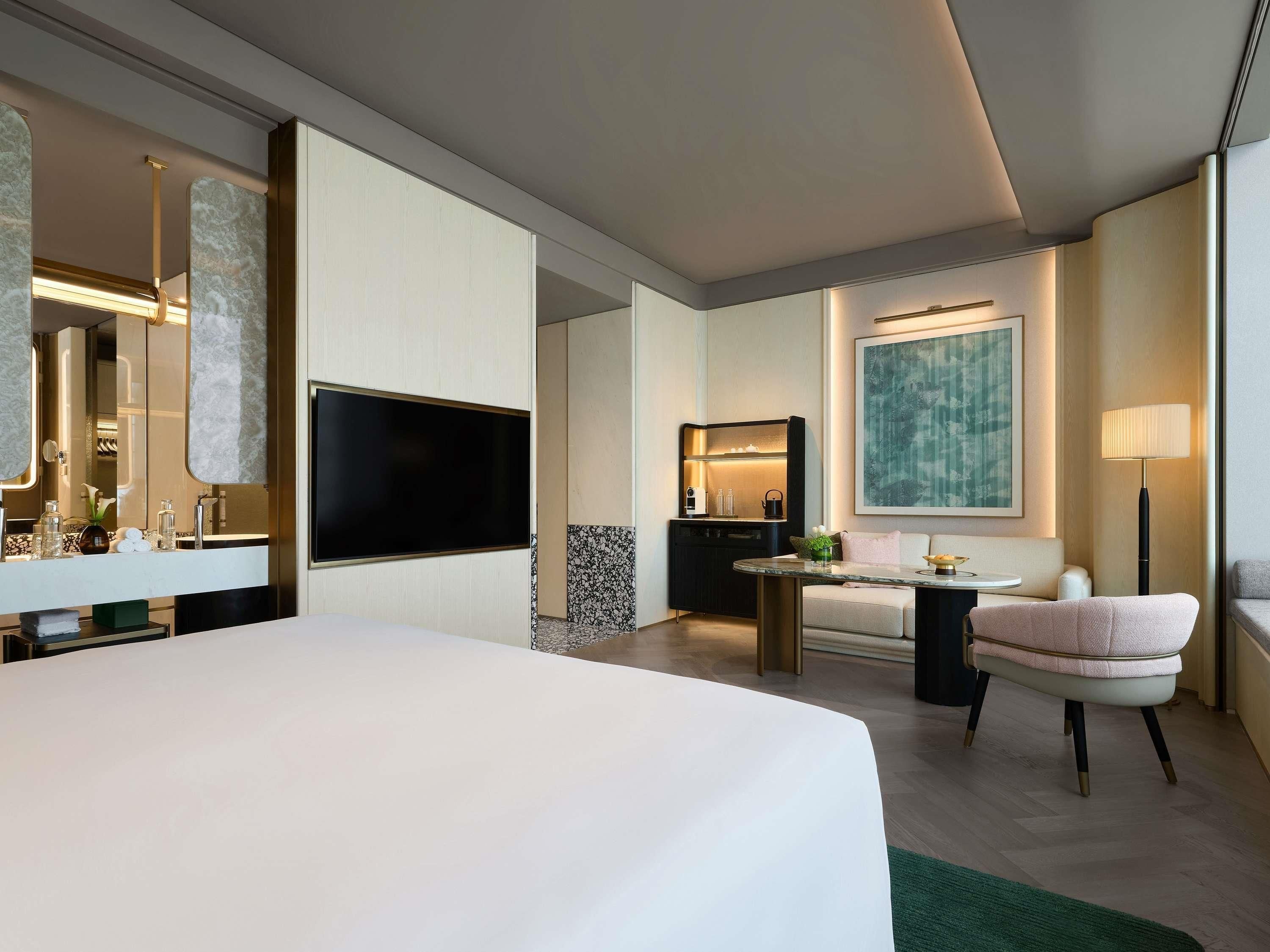 Une chambre d’hôtel moderne dotée d’un grand lit, d’un téléviseur à écran plat, d’un bureau avec une lampe, d’un canapé et d’un grand tableau abstrait accroché au mur.