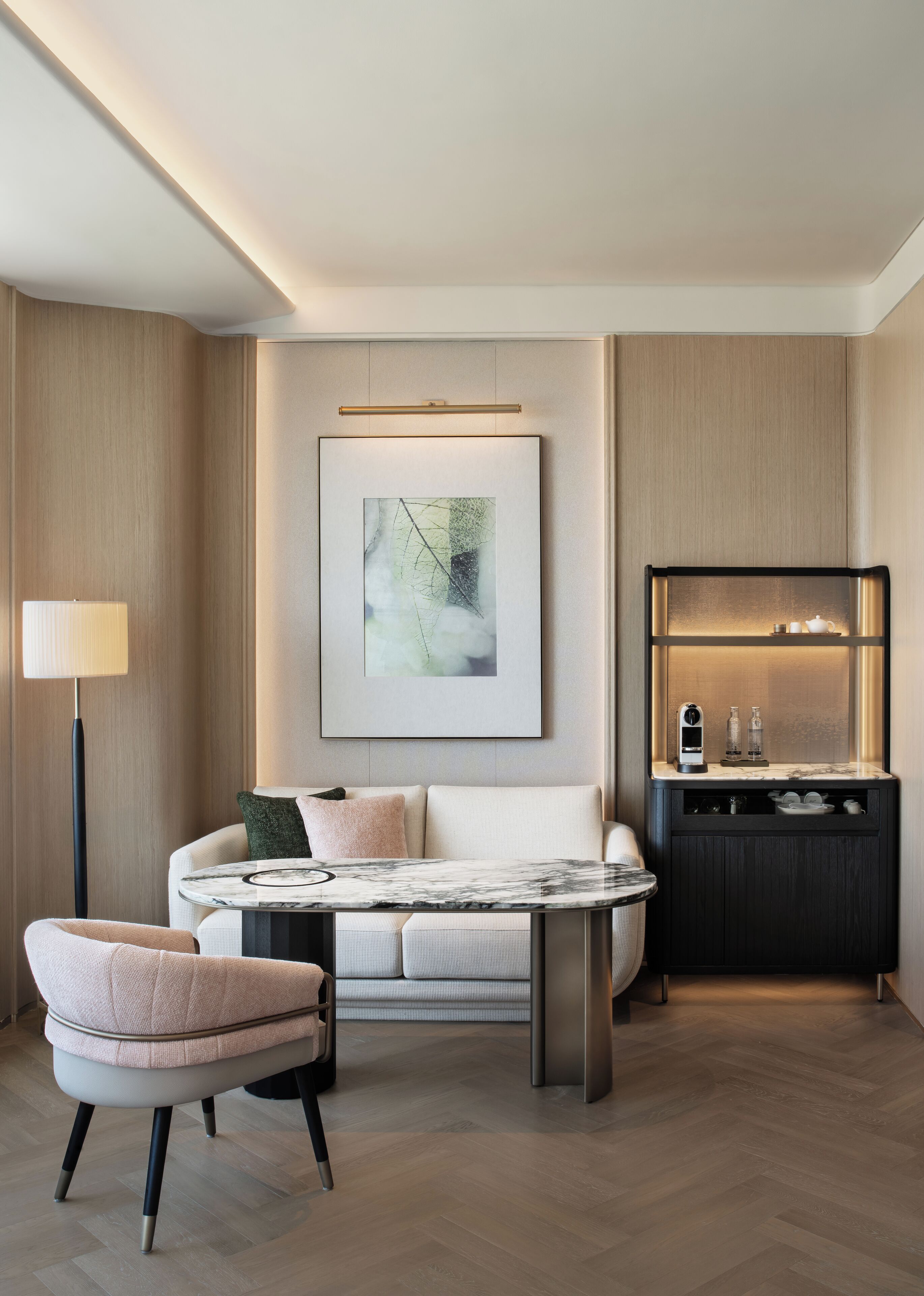 A modern hotel room with a sofa, a round table, a floor lamp, and a minibar.