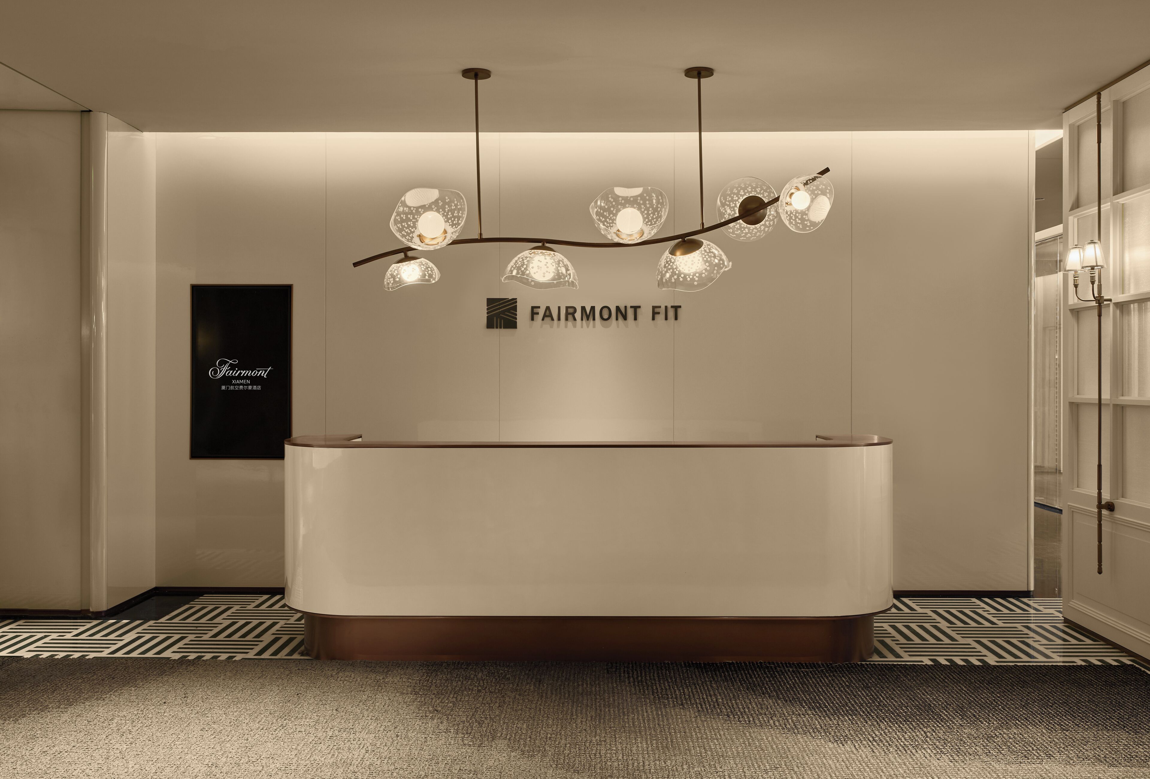 A modern reception area with a curved reception desk, a chandelier with multiple lights, and a sign that reads "FAIRMONT FIT".