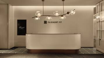 A modern reception area with a curved reception desk, a chandelier with multiple lights, and a sign that reads "FAIRMONT FIT".