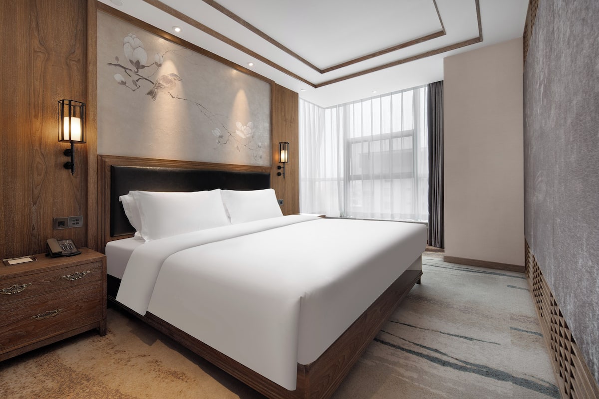 A modern hotel room with a large bed, wooden headboard, two bedside lamps, a nightstand, and a large window with sheer curtains.