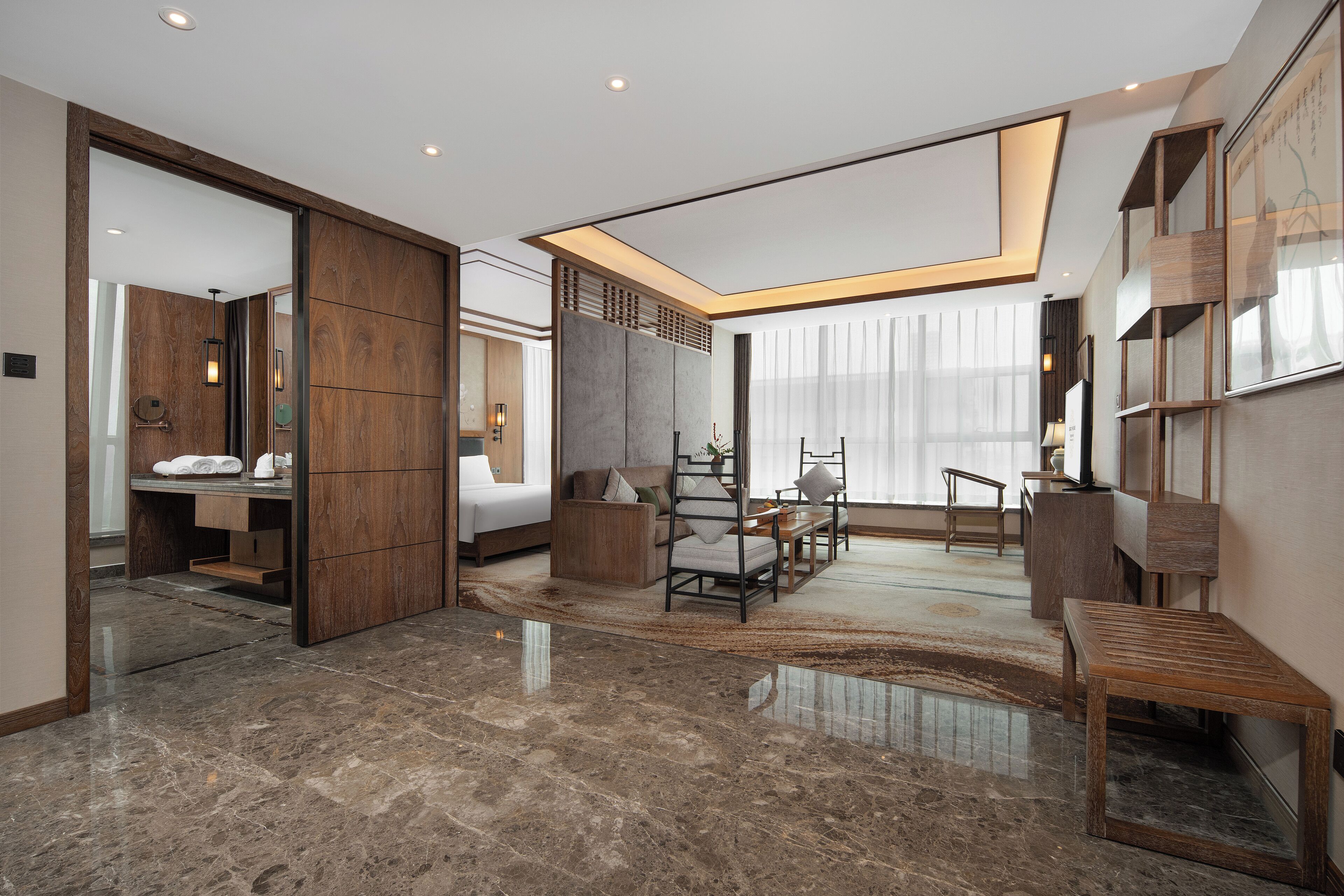 Une chambre d’hôtel moderne dotée d’un grand lit, d’un coin salon avec un canapé et des fauteuils, d’un buffet en bois et d’une grande fenêtre ornée de rideaux légers.