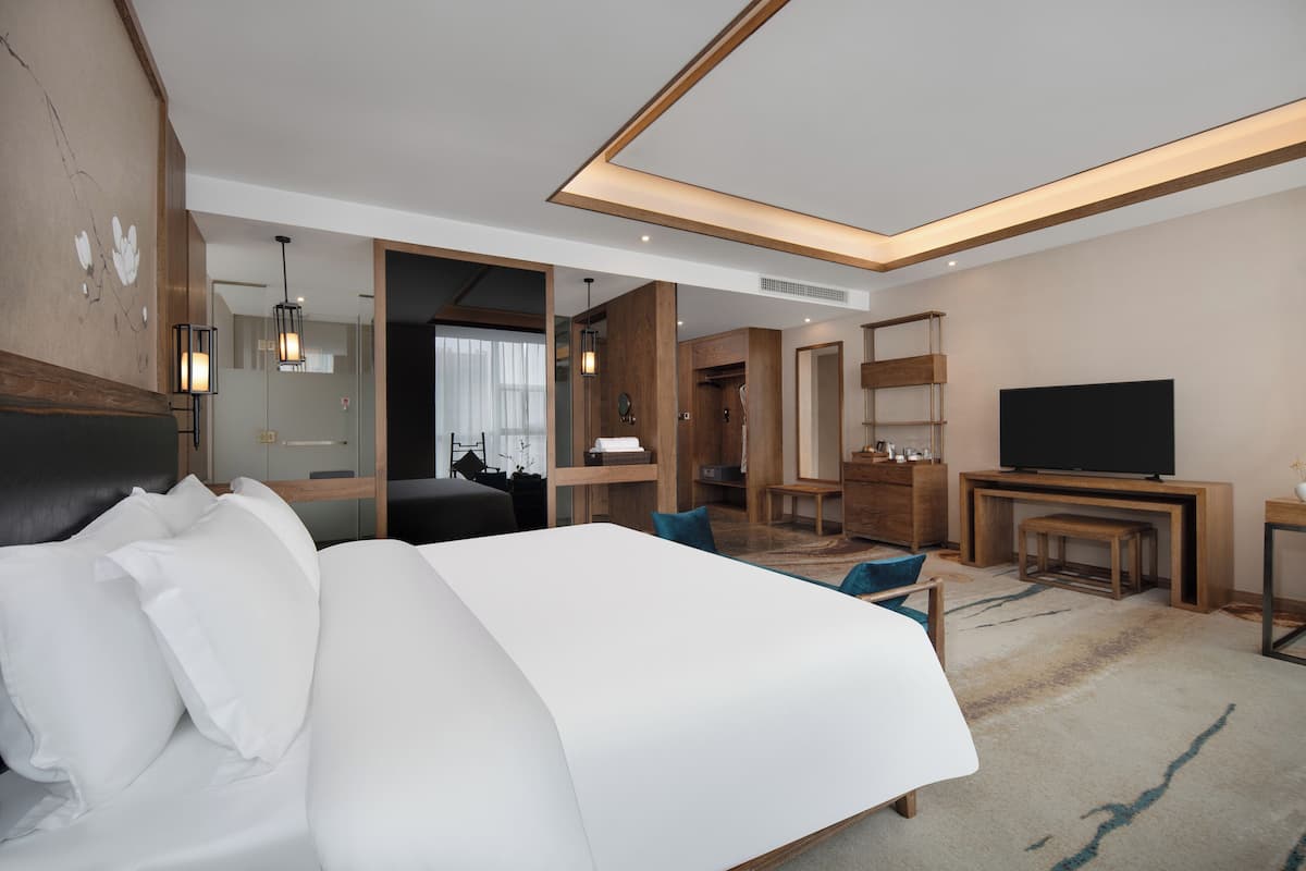 A modern hotel room with a large bed, a TV, a desk, and a bathroom visible through an open door.