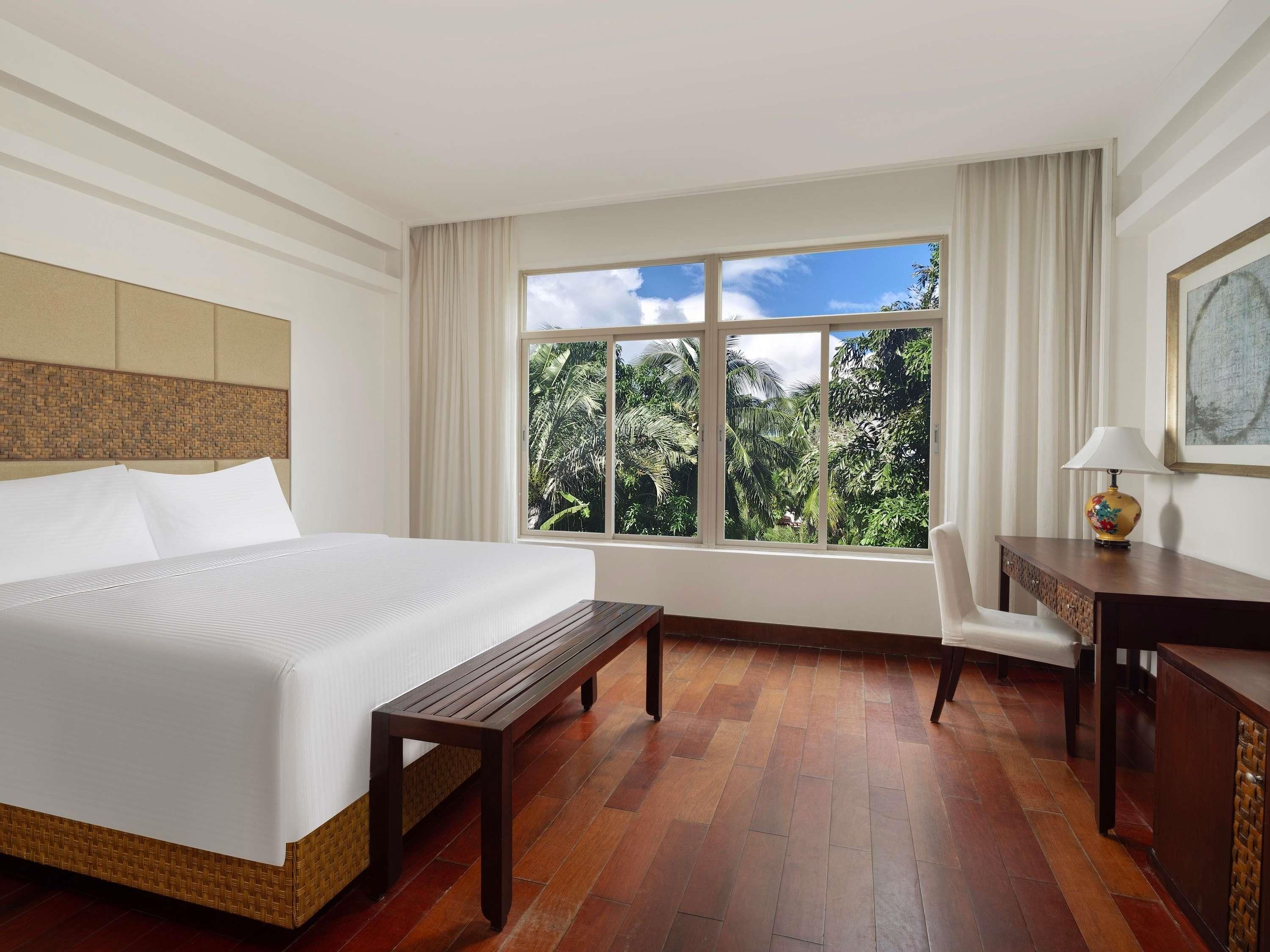 A hotel room with a large bed, a wooden bench, a desk, and a window with a view of greenery.