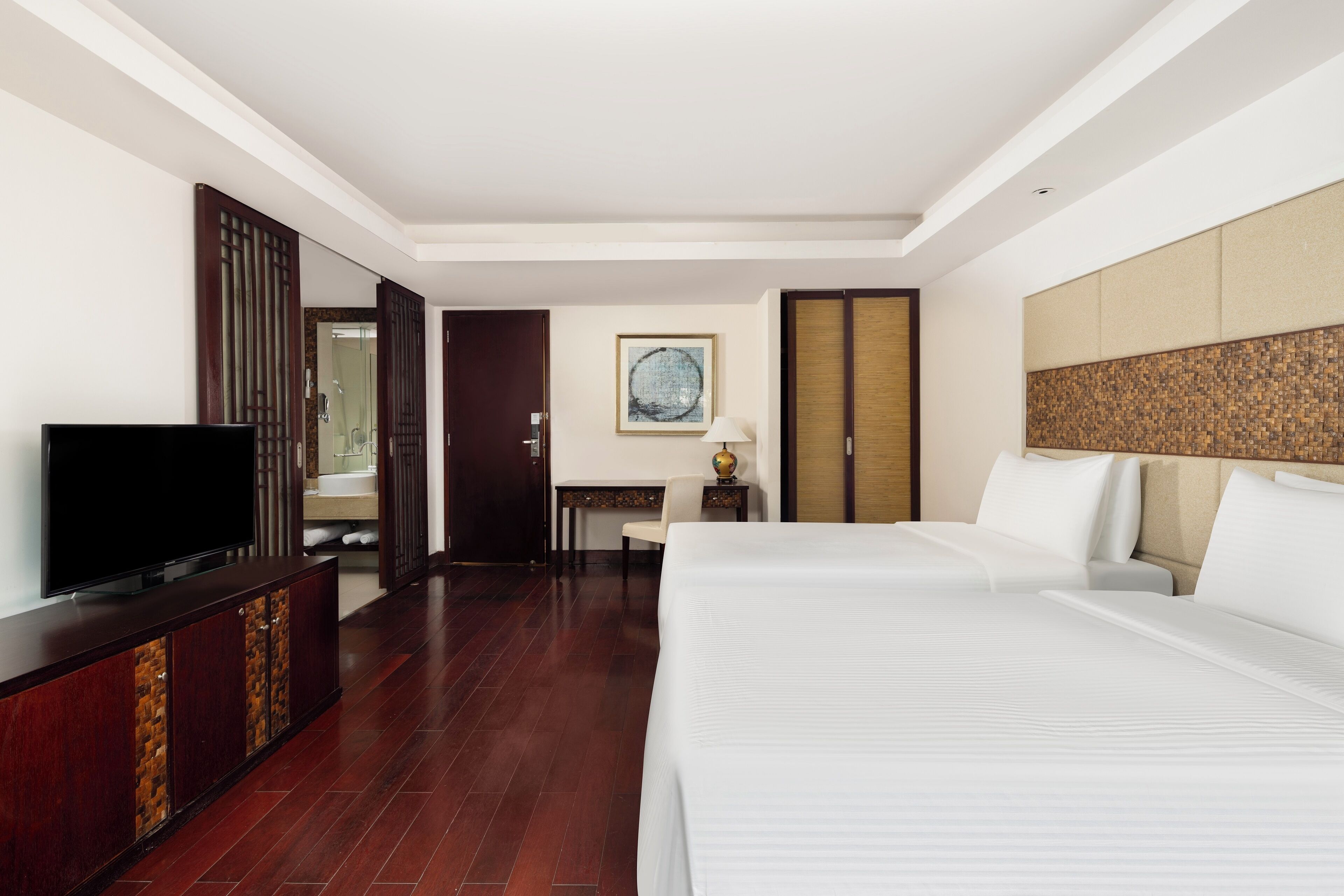 A hotel room with a large bed, a television on a wooden cabinet, a desk with a lamp, and a view of the bathroom.