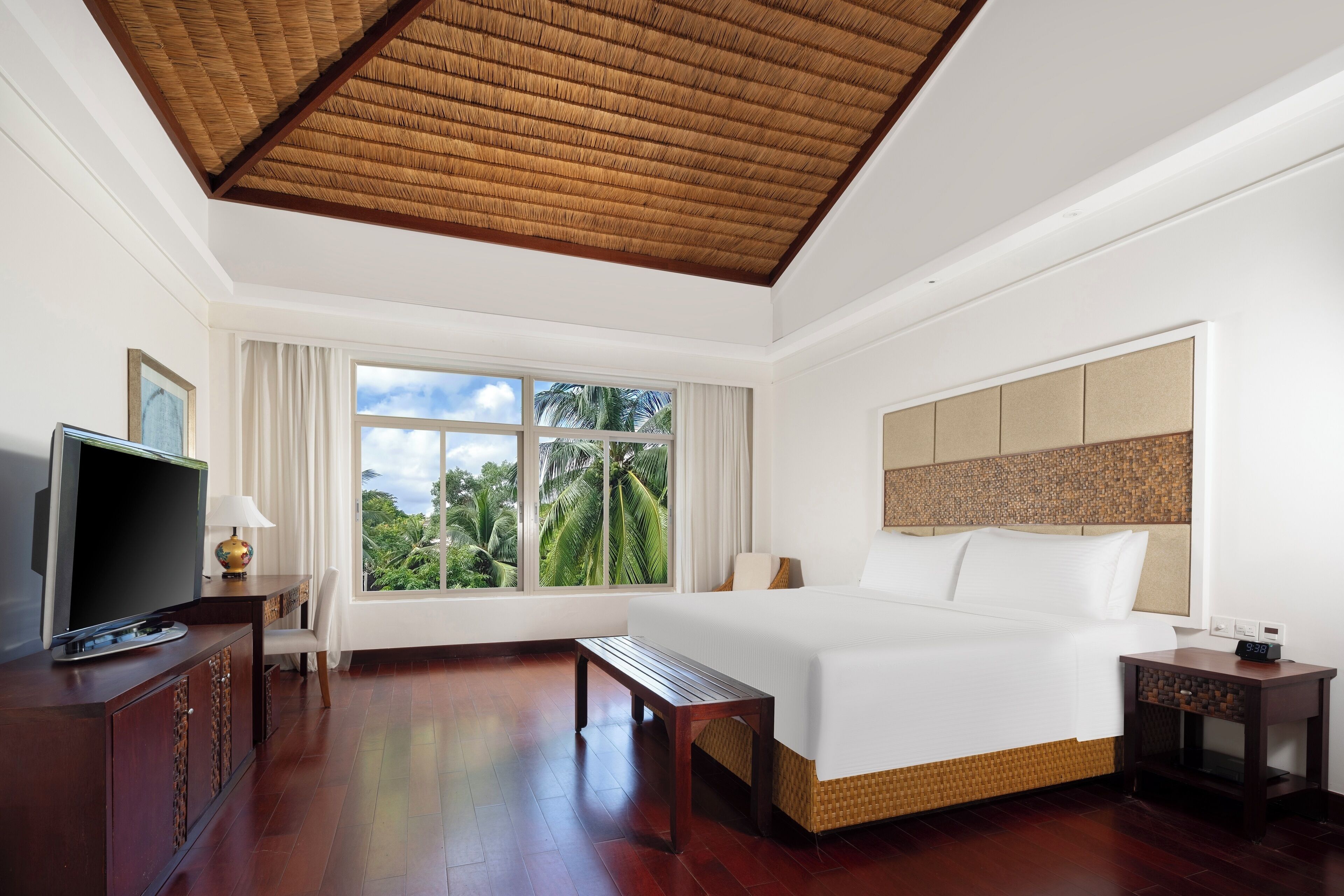 A hotel room with a large bed, a television, a desk, and a window with a view of palm trees.