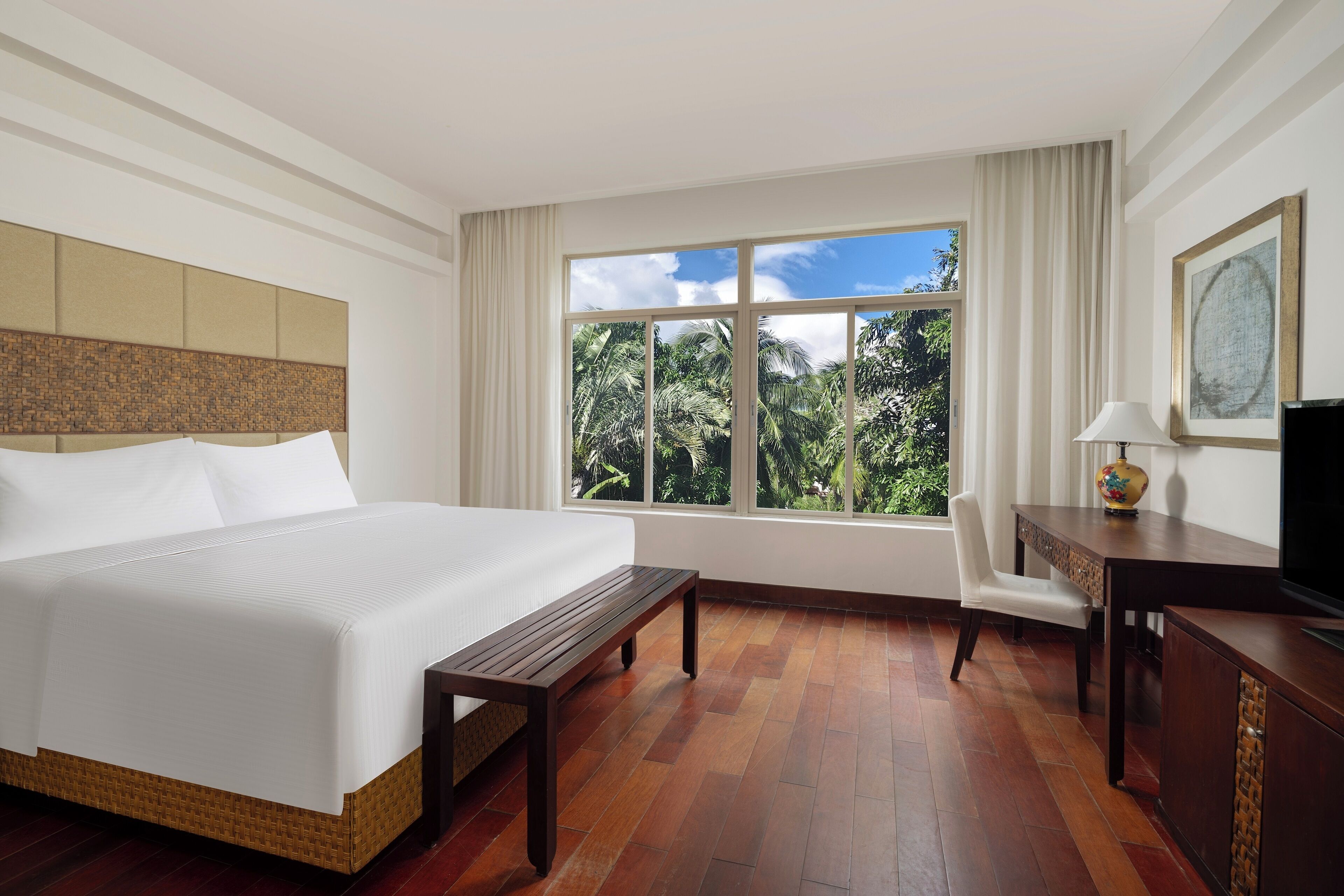 A hotel room with a large bed, a wooden bench, a desk with a lamp, and a view of palm trees through a window.