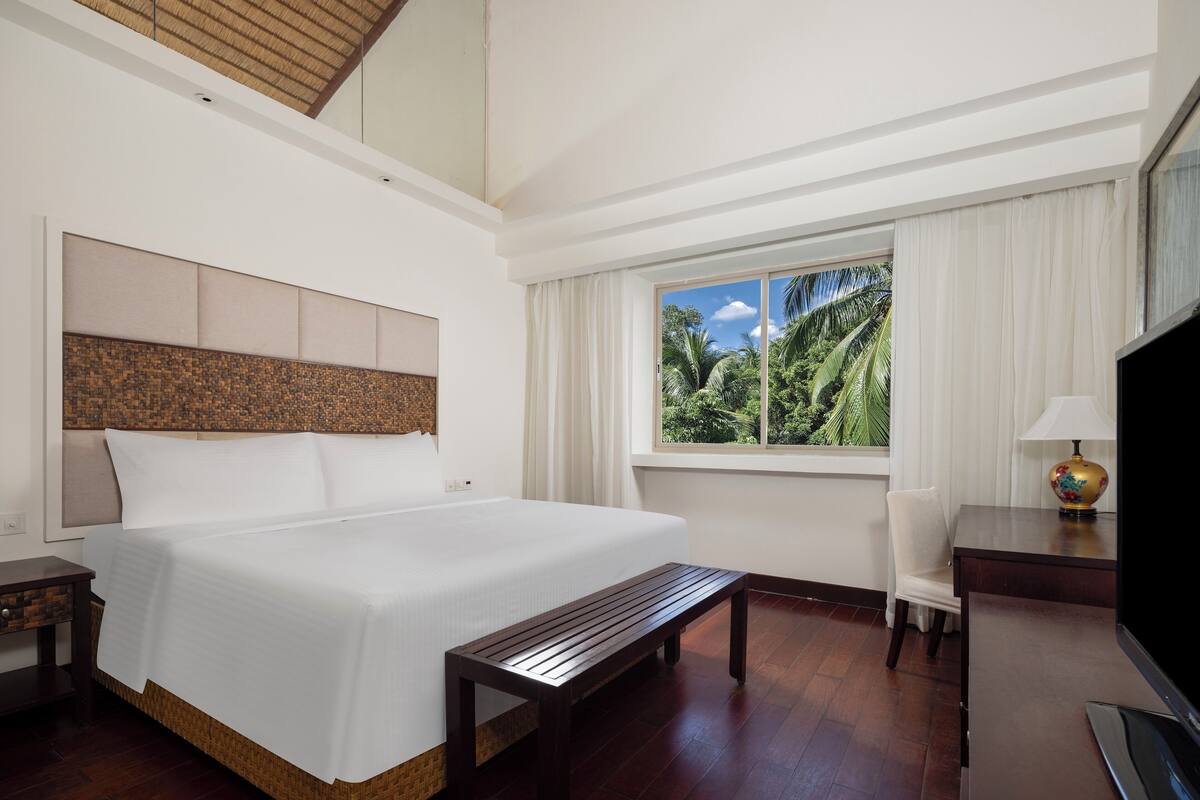 A hotel room with a large bed, a wooden bench, a desk with a lamp, and a window with a view of palm trees.