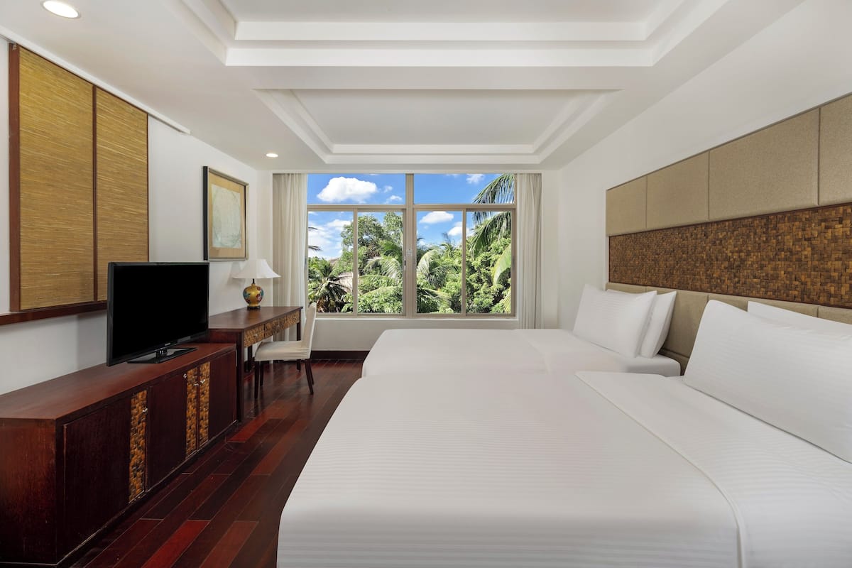 A hotel room with a large bed, a TV on a wooden cabinet, a desk with a lamp, and a view of greenery through the window.