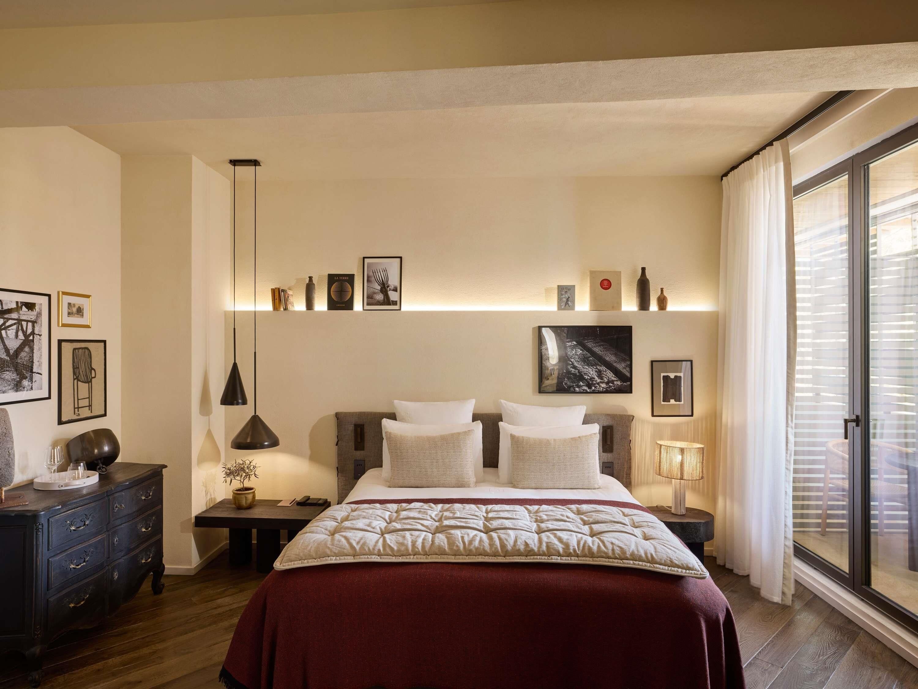 Quarto com cama, mesas de cabeceira, cómoda, janela com persianas e estantes na parede com quadros e objetos decorativos.
