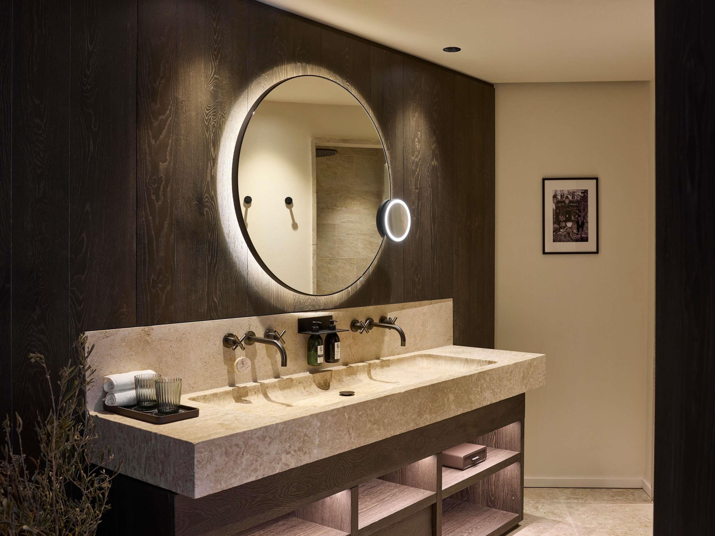A modern bathroom with a large vanity, a round mirror with a light, and a framed picture on the wall.