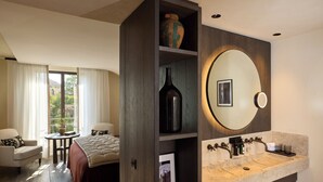A modern hotel room with a large bed, a sitting area with a table and chairs, a built-in vanity with a mirror, and a view through a large window. - L'Isle de Leos Hotel & Spa - MGallery Collection (L'Isle-sur-la-Sorgue)