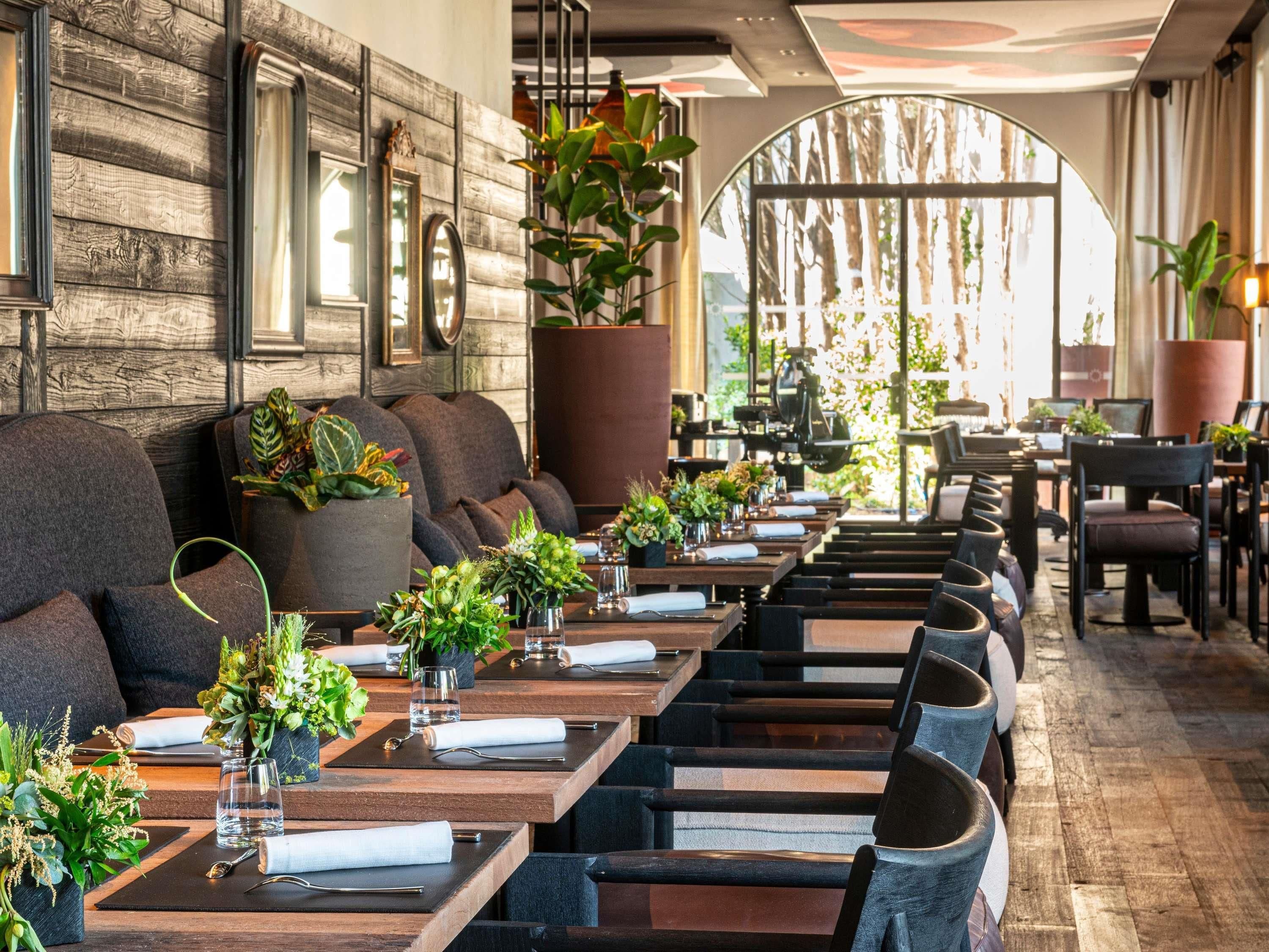 A restaurant with long wooden tables, dark upholstered chairs, and potted plants.