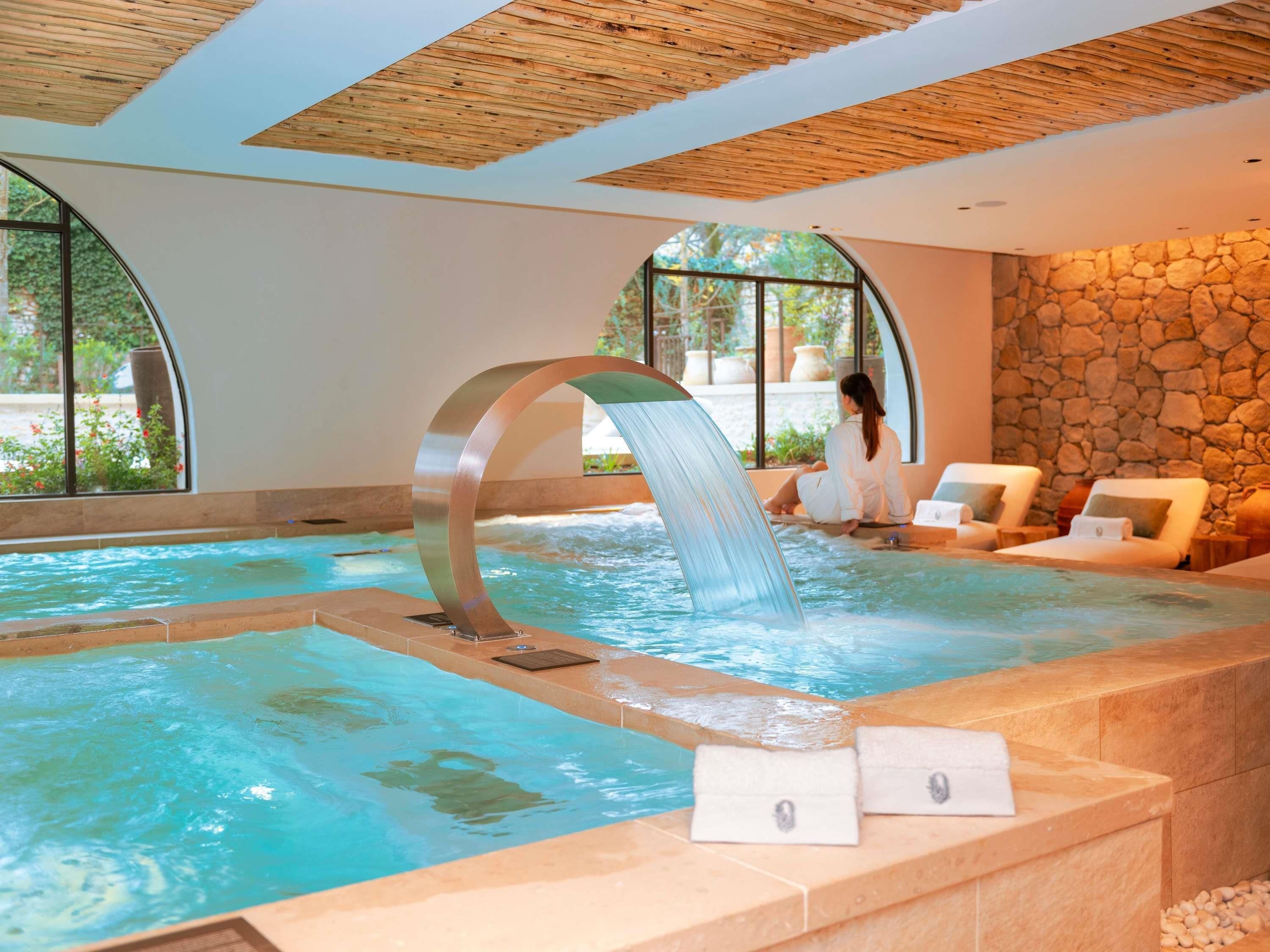 A spa pool with a waterfall feature, lounge chairs, and a stone wall.