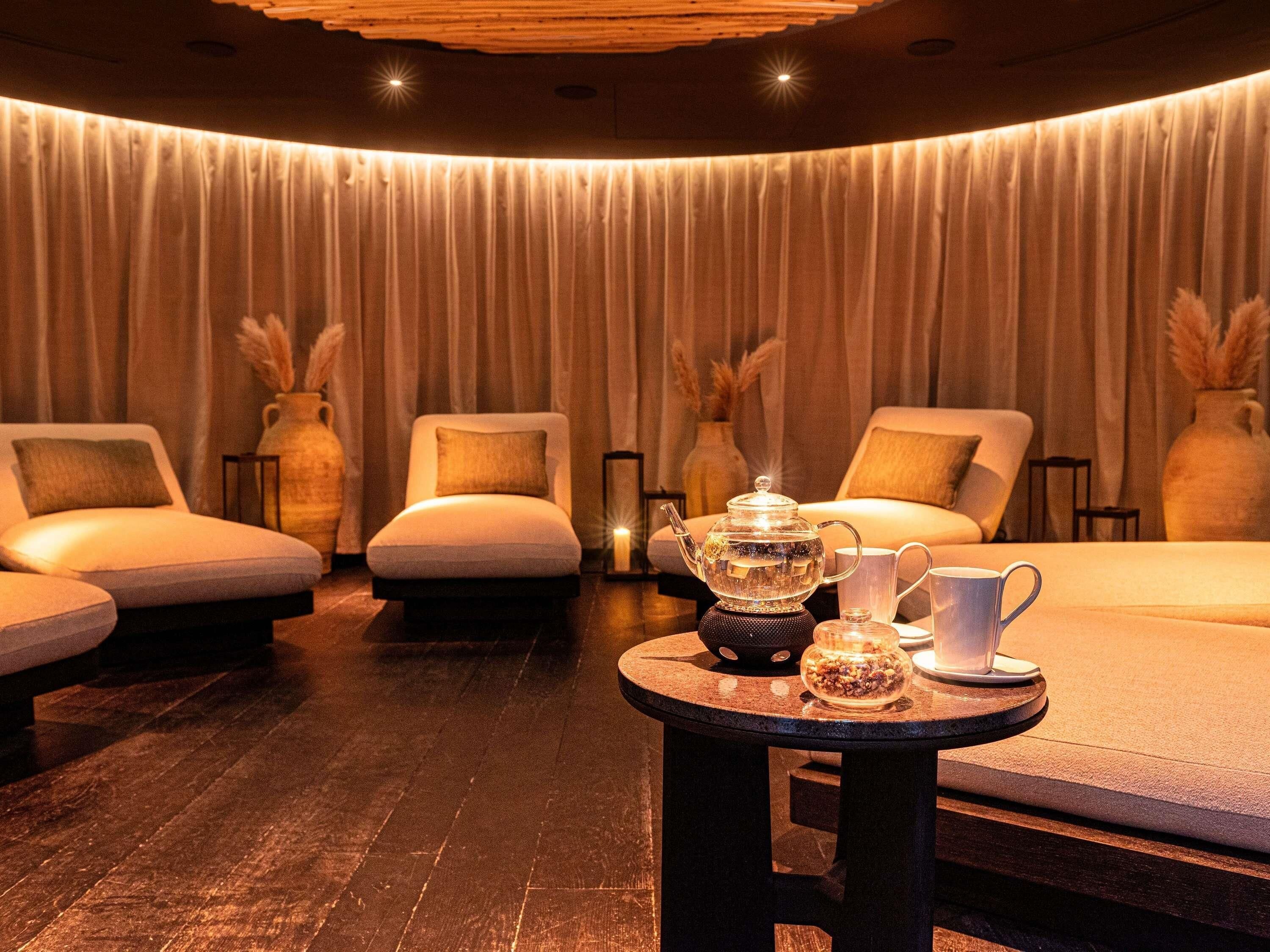 Ein Spa-Behandlungsraum mit Massagetisch, zwei Stühlen, einem Tisch mit Teekanne und Tassen sowie einem Vorhang im Hintergrund.