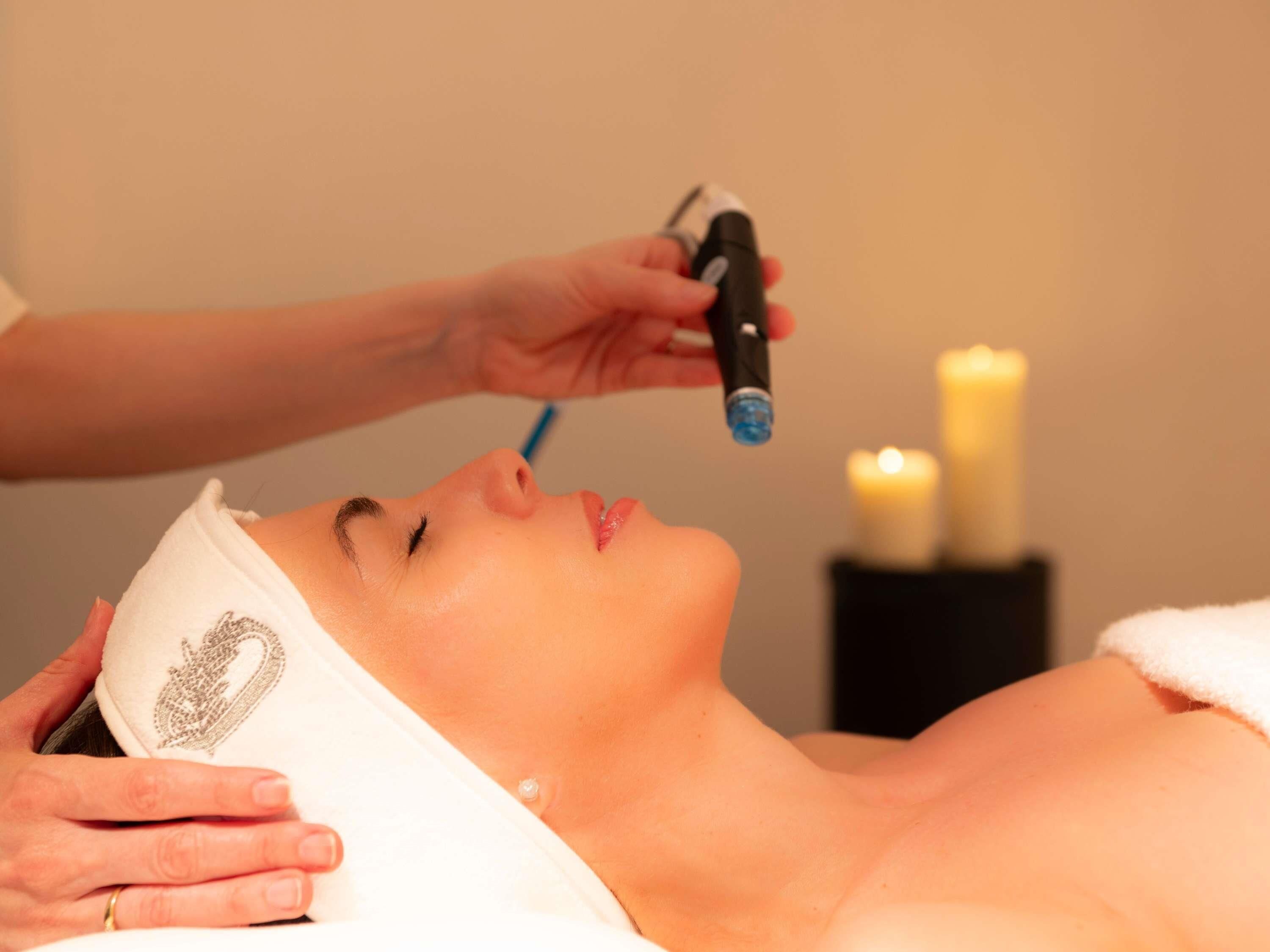 A person receiving a facial treatment with a device, a white towel, and candles in the background.