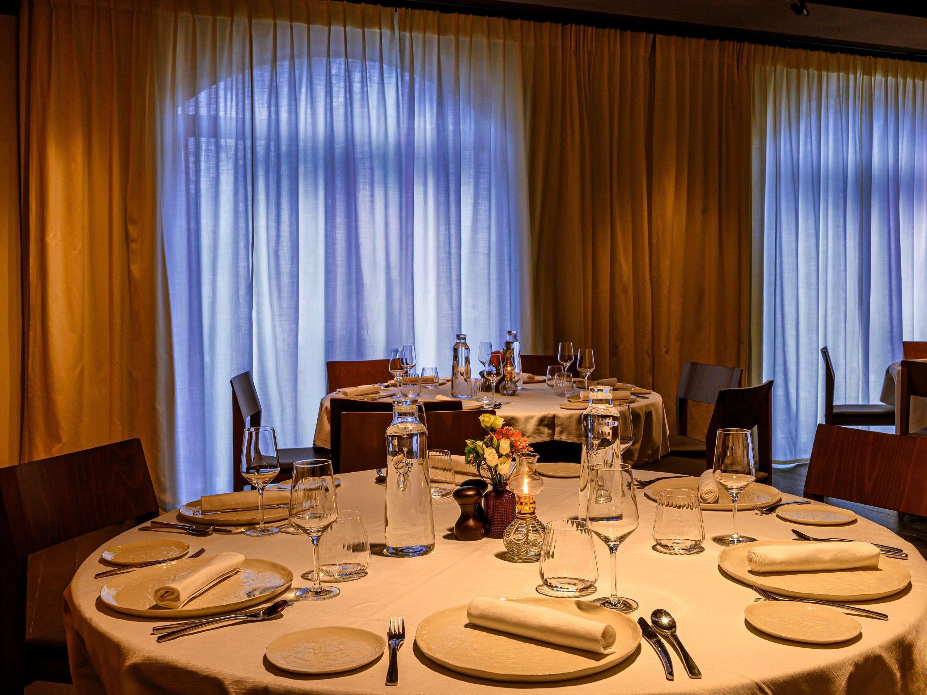 Comedor con mesas redondas dispuestas para dos, con manteles blancos, servilletas dobladas con esmero y copas de vino vacías.