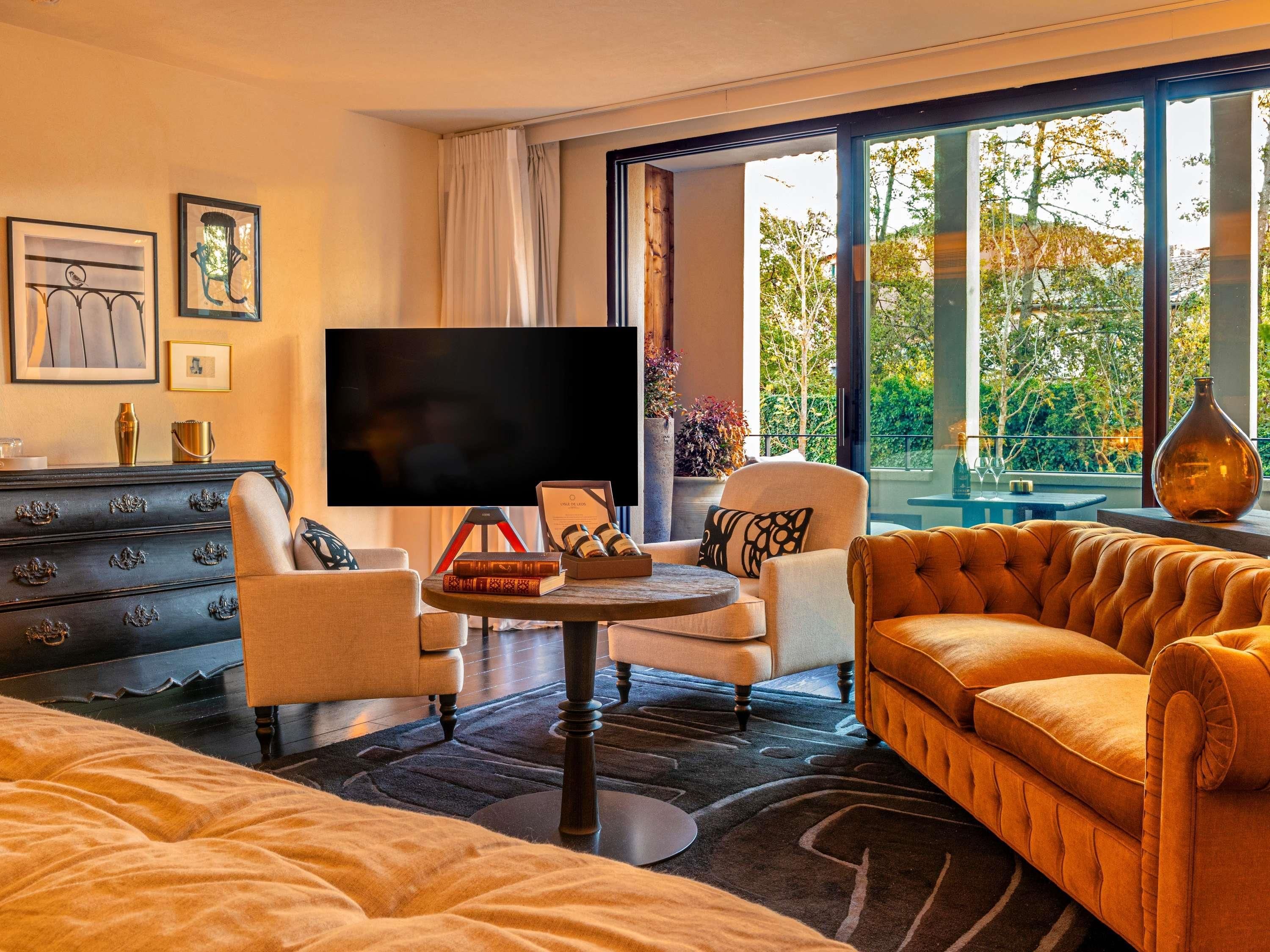 Une chambre d’hôtel moderne dotée d’une grande fenêtre, d’un téléviseur à écran plat, d’un canapé, d’un fauteuil, d’une petite table et d’une commode.