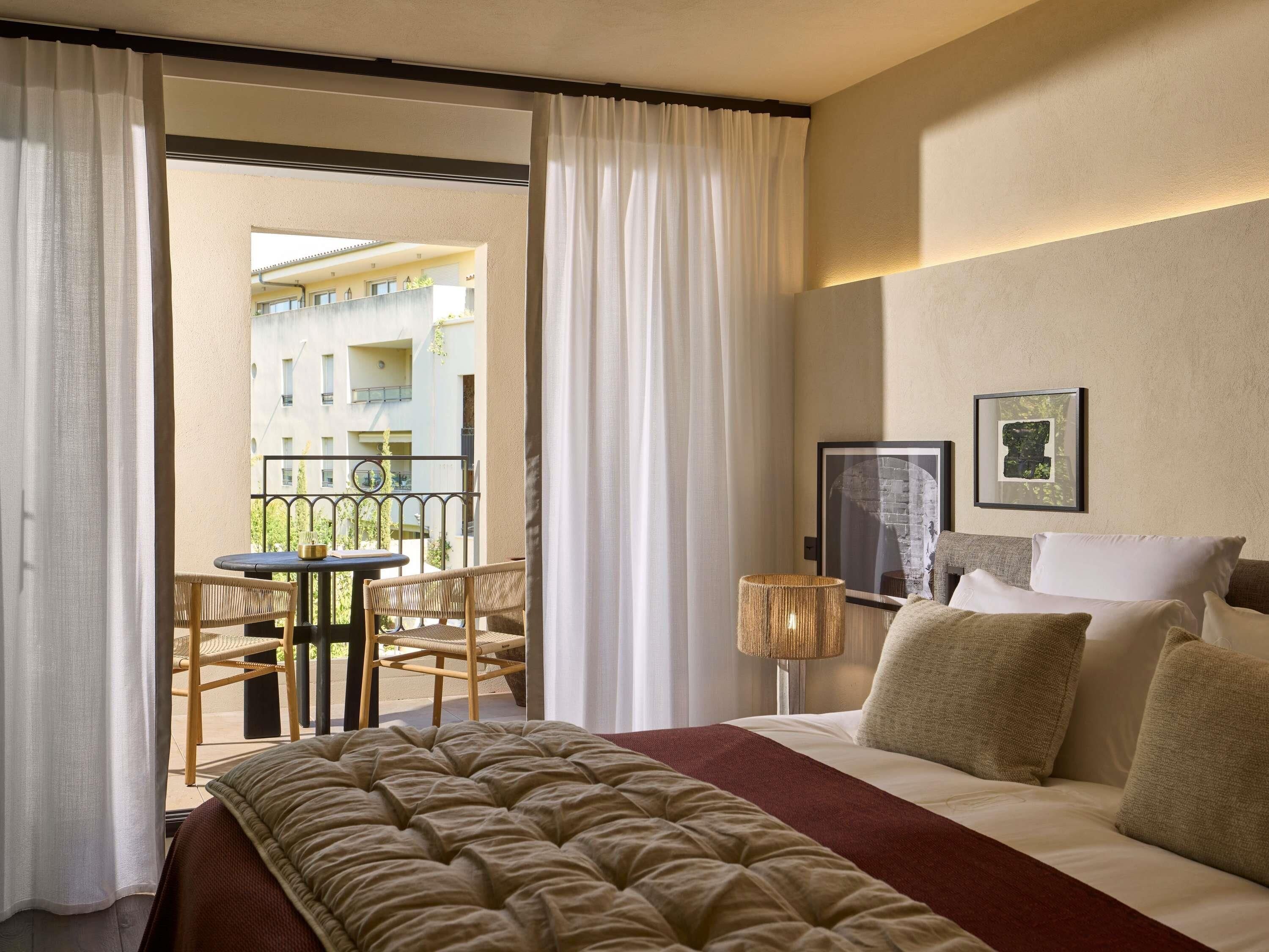 A hotel room with a large bed, a small table with chairs, a lamp, and a balcony with a view of buildings.
