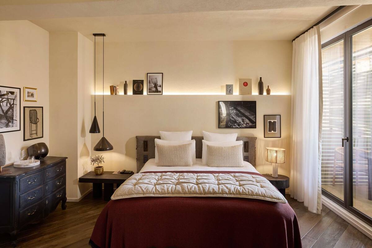 A bedroom with a bed, bedside tables, a dresser, a window with blinds, and wall-mounted shelves with pictures and decorative items.