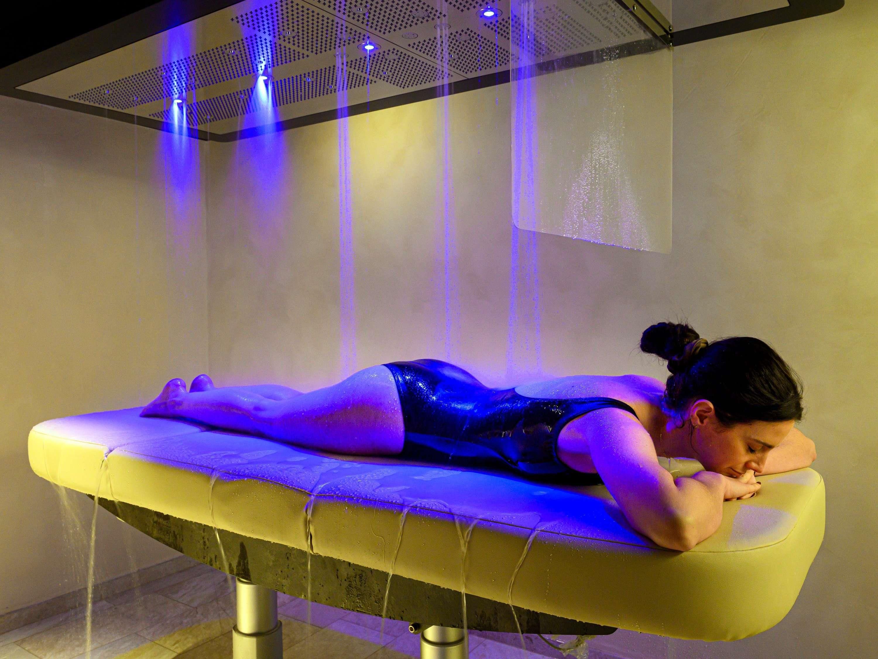 A person lying on a massage table with water jets, in a room with a ceiling grid and a light fixture.