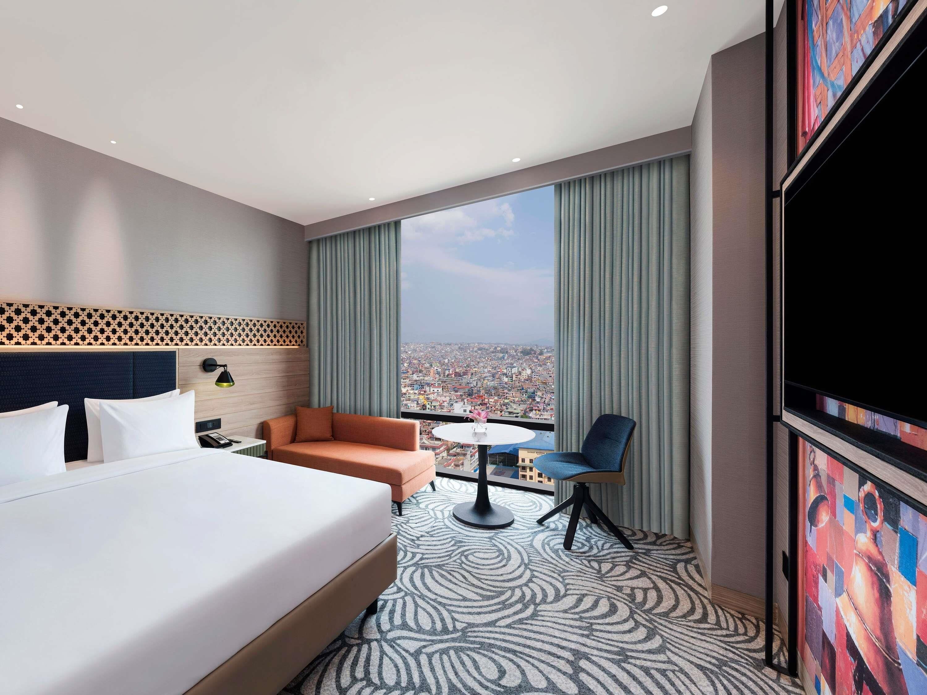 A hotel room with a bed, a sofa, a chair, a small table, and a large window with a city view.