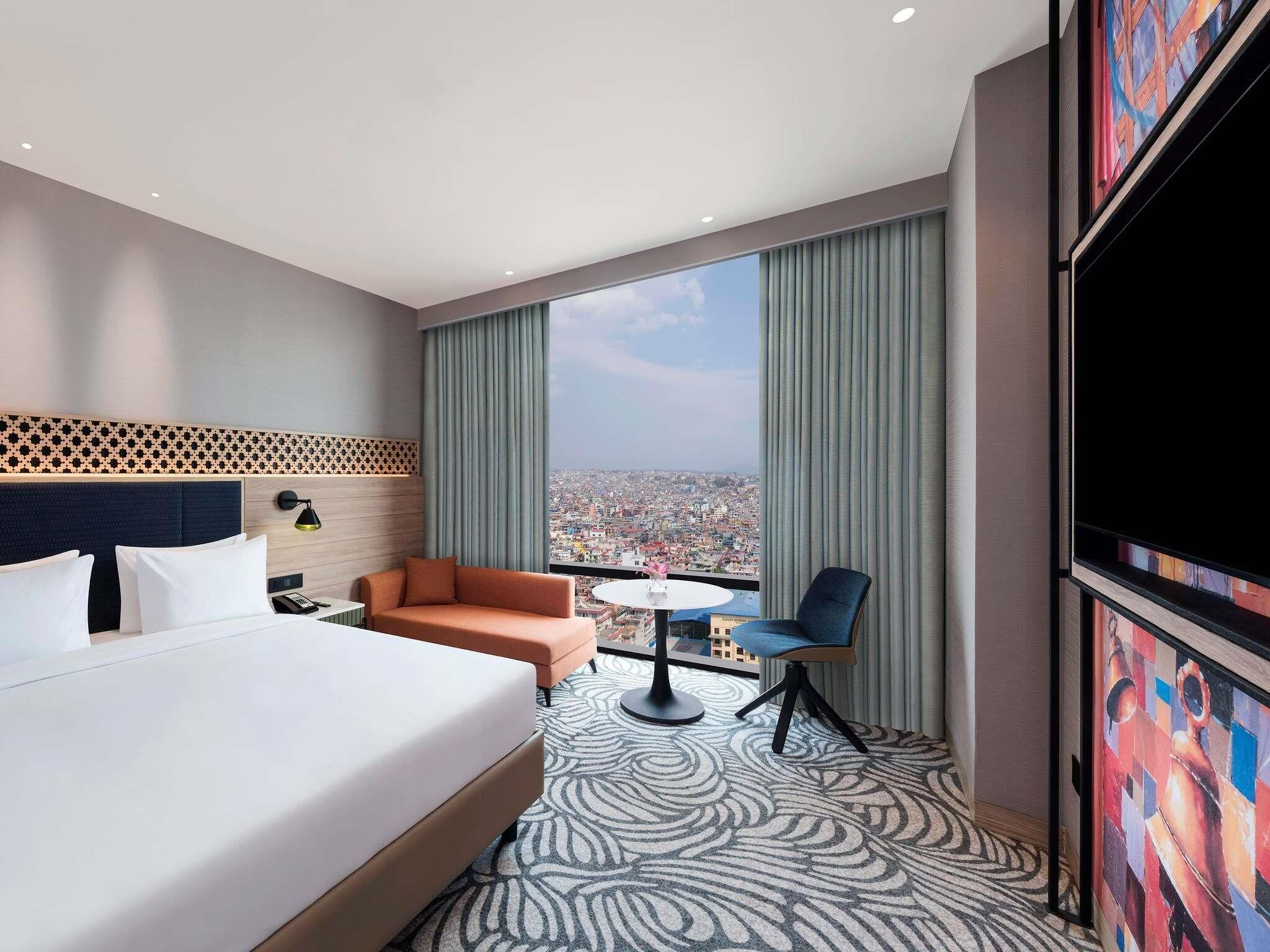A hotel room with a large bed, a sofa, a small table, a chair, and a large window with a city view.