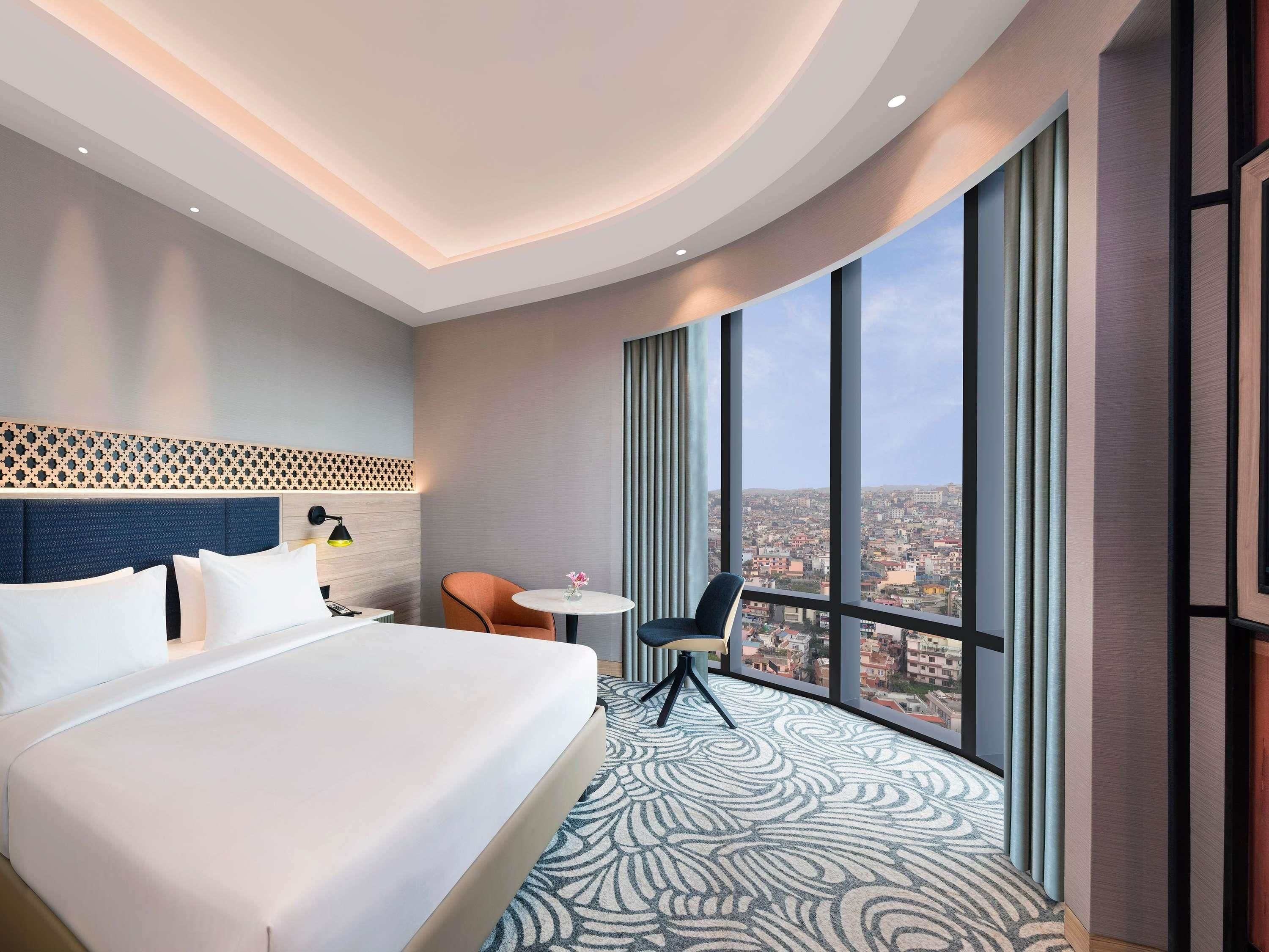 A hotel room with a large bed, a small table, a chair, and a view of the city through large windows.