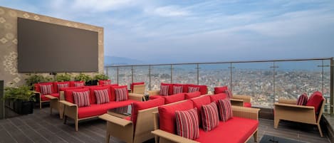 A rooftop terrace with wicker furniture and red cushions, offering a panoramic city view.