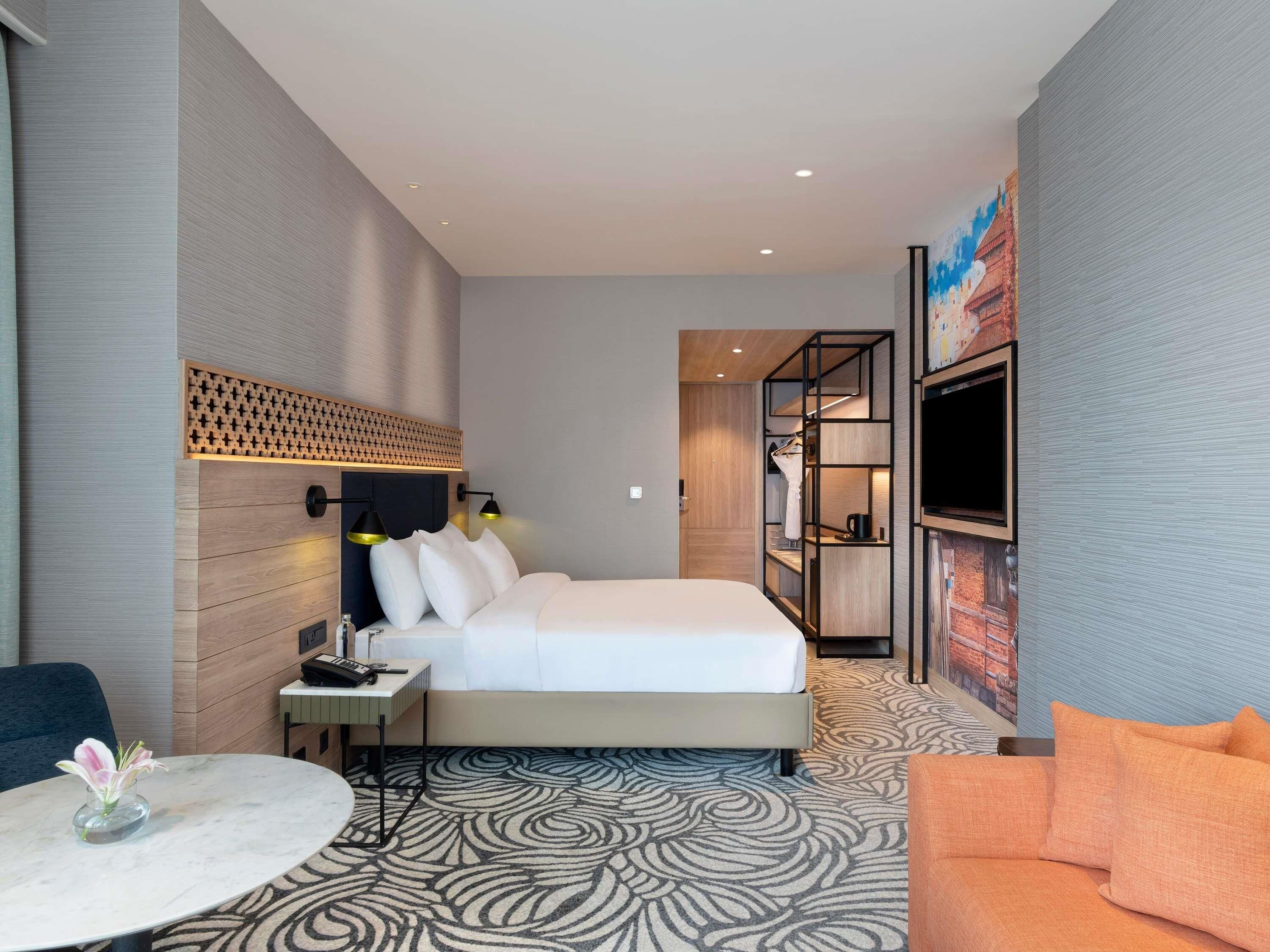 Quarto de hotel moderno com cama, mesa de cabeceira, luminária, televisão e carpete estampado.