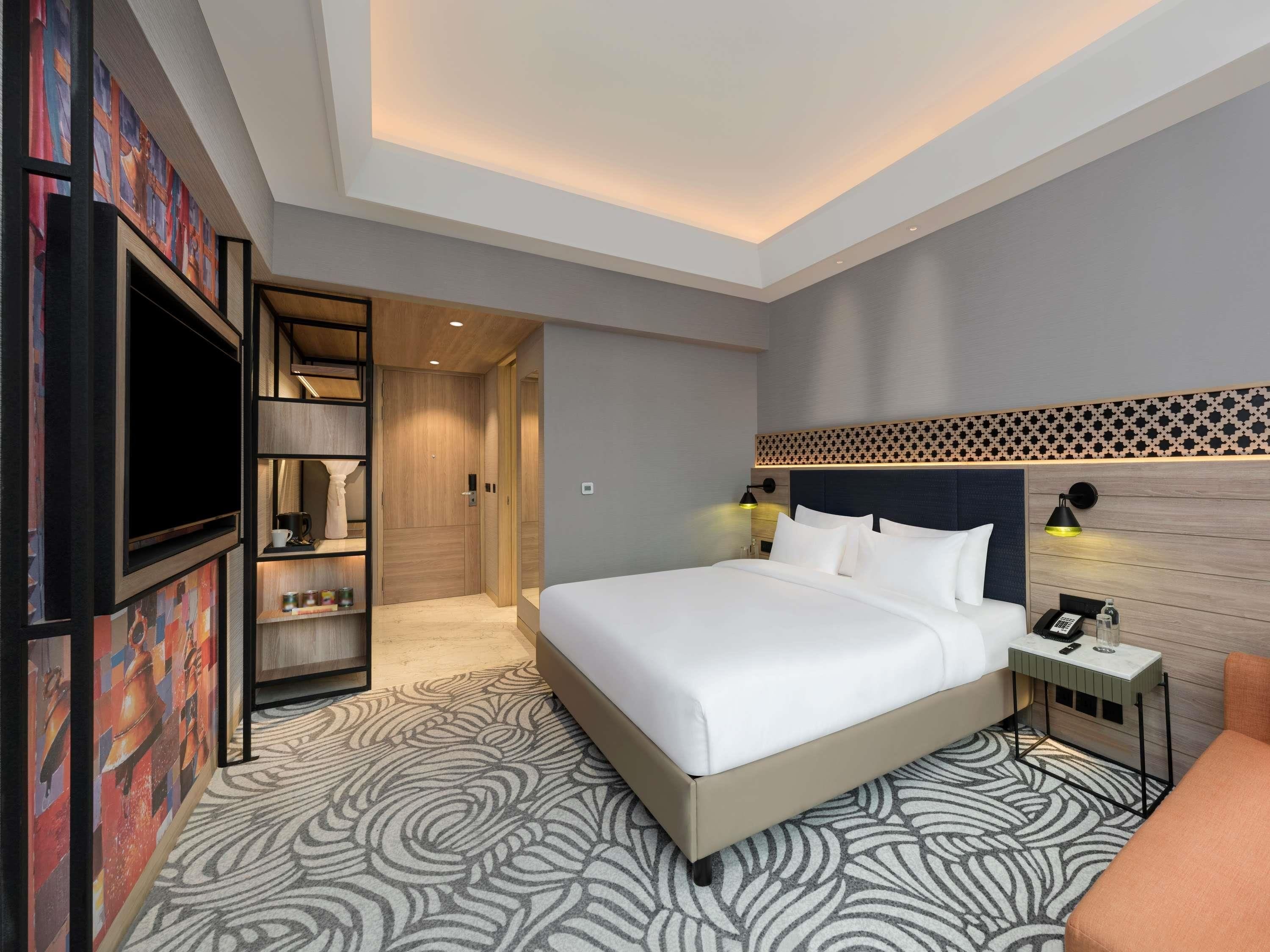 A modern hotel room with a large bed, a TV, a bedside table with a lamp, and a small seating area.
