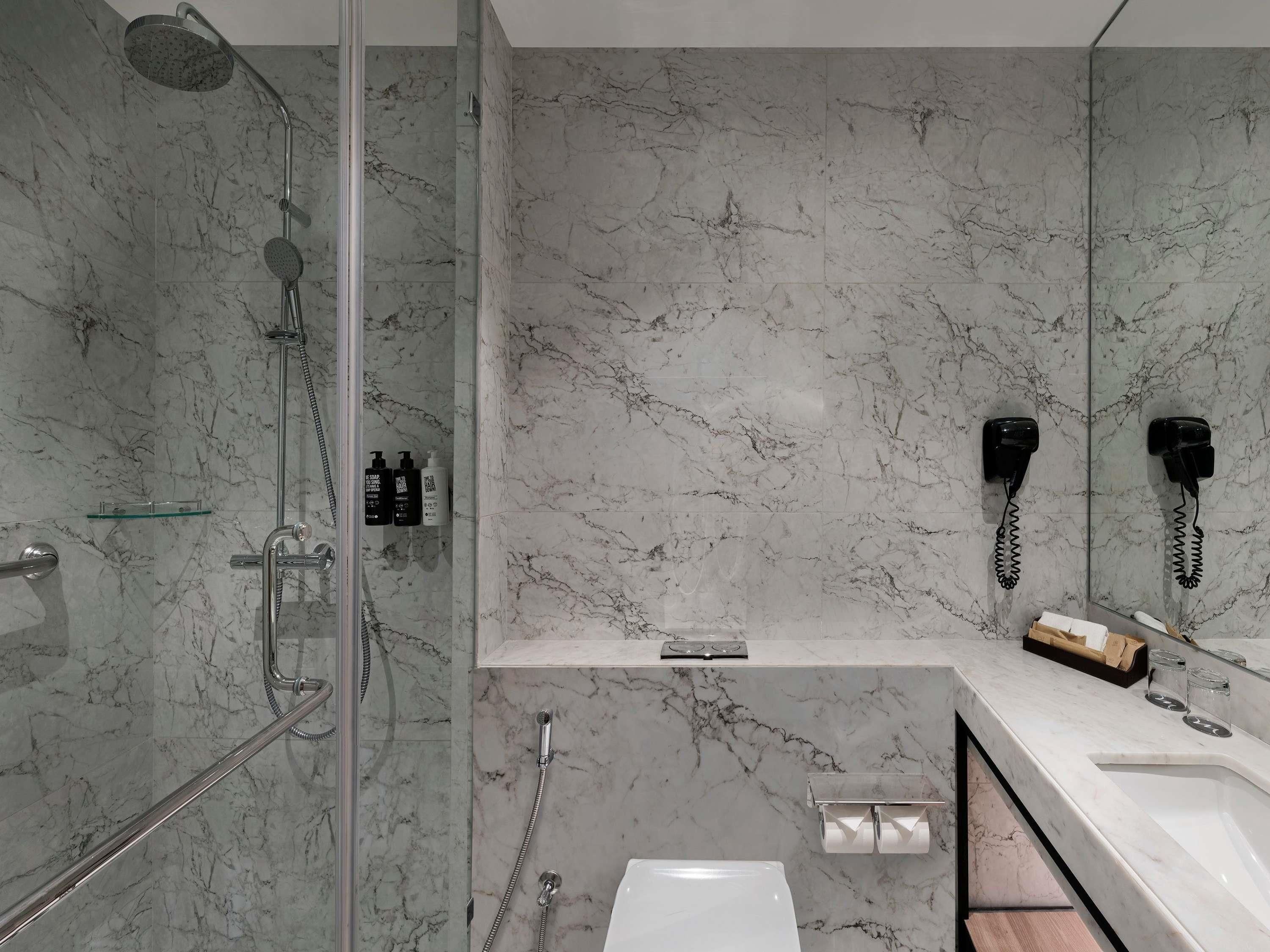 Une salle de bain moderne dotée d’une douche à parois de verre, d’un mur recouvert de carreaux de marbre, d’un lavabo blanc et d’un miroir avec un téléphone intégré.