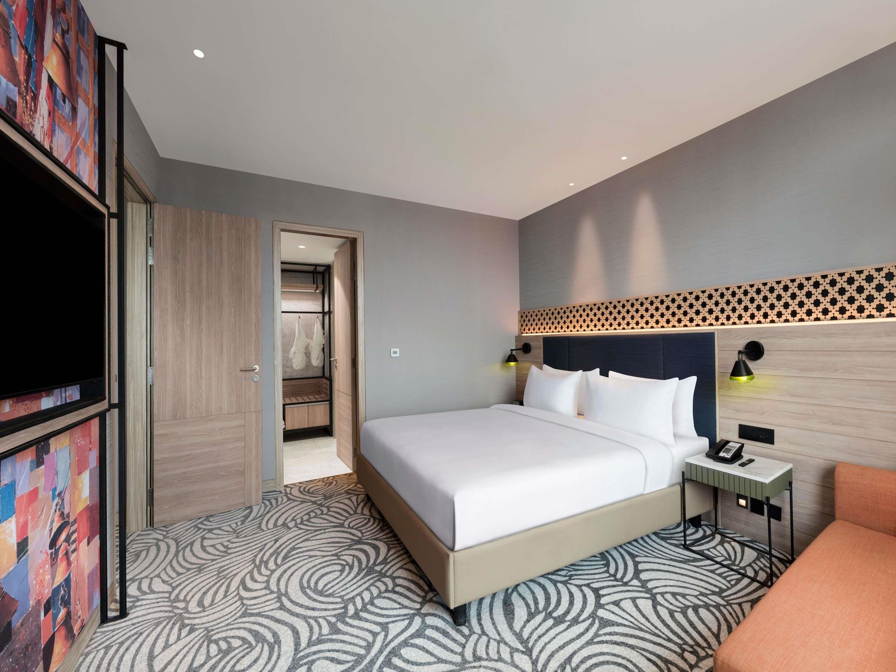 A modern hotel room with a large bed, a bedside table with a lamp, a TV, and a wardrobe.