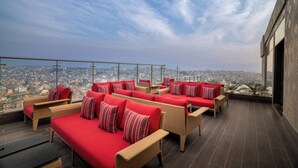 Terraço no telhado com móveis de vime e almofadas vermelhas, proporcionando vista da cidade.