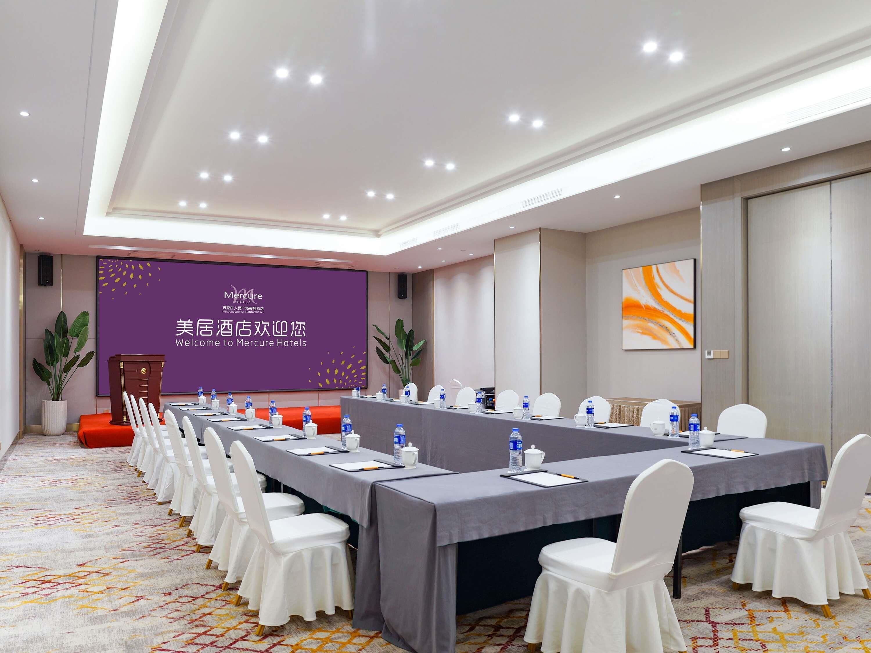 A conference room with a long table, white chairs, and a presentation screen.