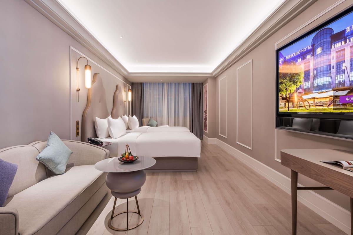 A modern hotel room with a large bed, a sofa, a round table, and a TV displaying a hotel exterior.