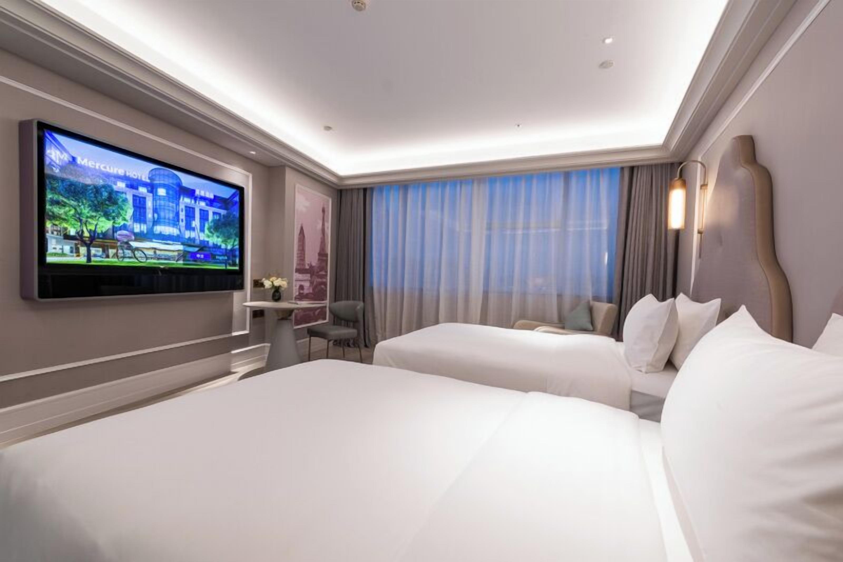 A hotel room with two beds, a TV mounted on the wall, a desk with a chair, and a window with curtains.