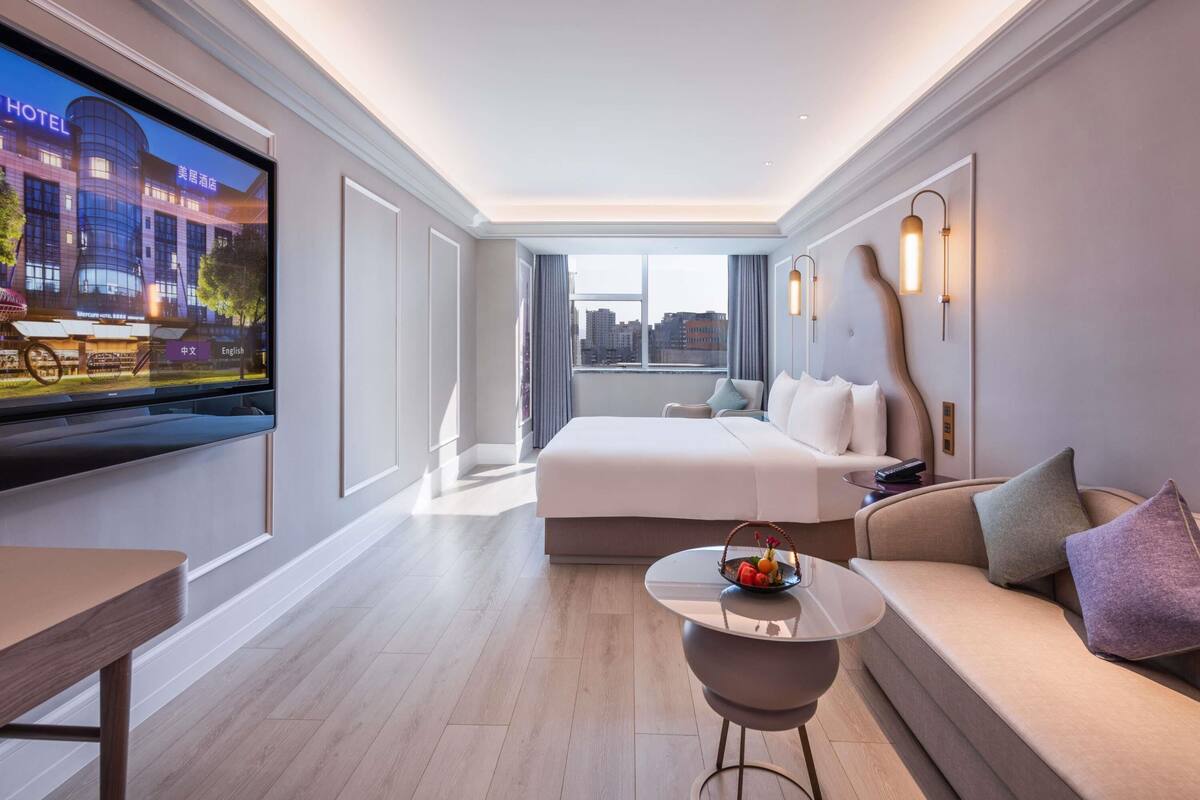 A modern hotel room with a large bed, a TV, a small table with fruit, and a sofa.