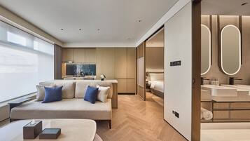Una habitación de hotel moderna con sofá, mesitas de noche, escritorio, ventana grande y baño con tocador y espejo.