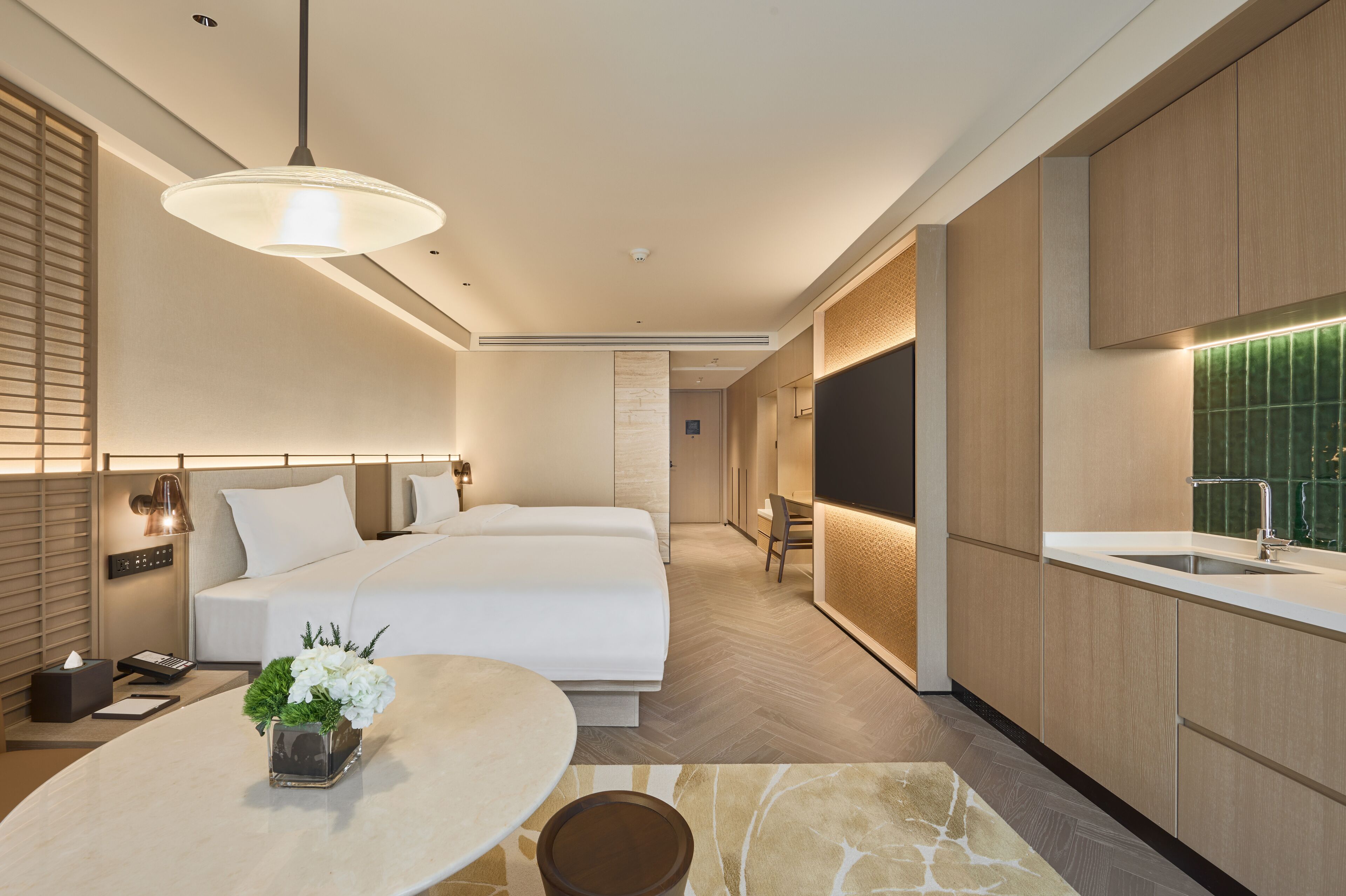 A modern hotel room with a large bed, a dining area with a table and chairs, a kitchen with a sink, and a bathroom with a shower.