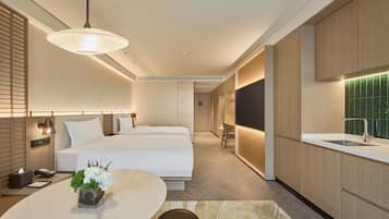 A modern hotel room with a large bed, a dining area with a table and chairs, a kitchen with a sink, and a bathroom with a shower.