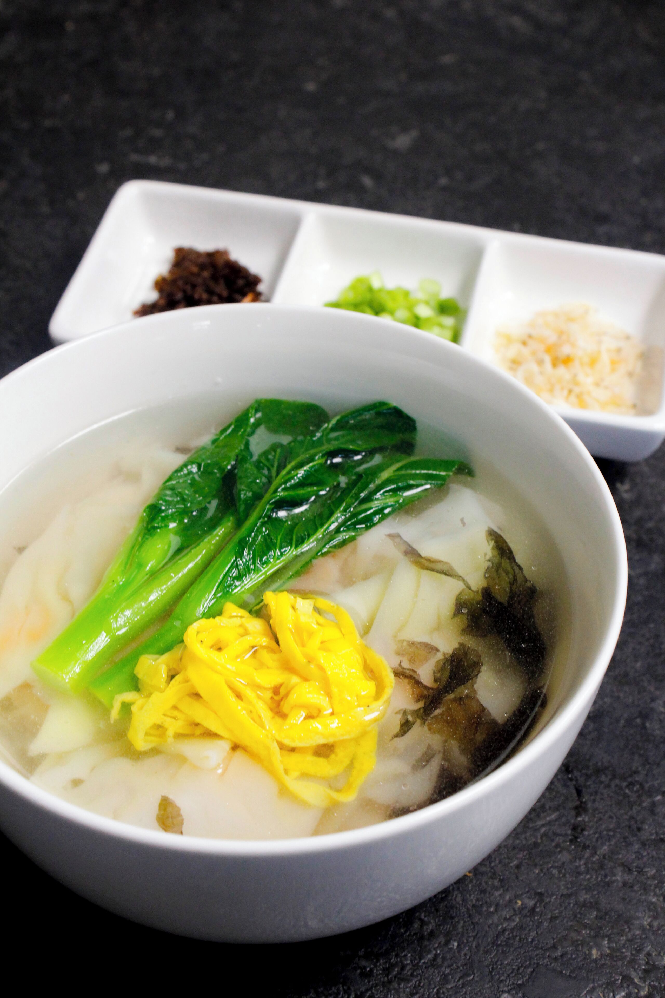 A bowl of soup with vegetables and noodles, accompanied by small dishes of condiments.