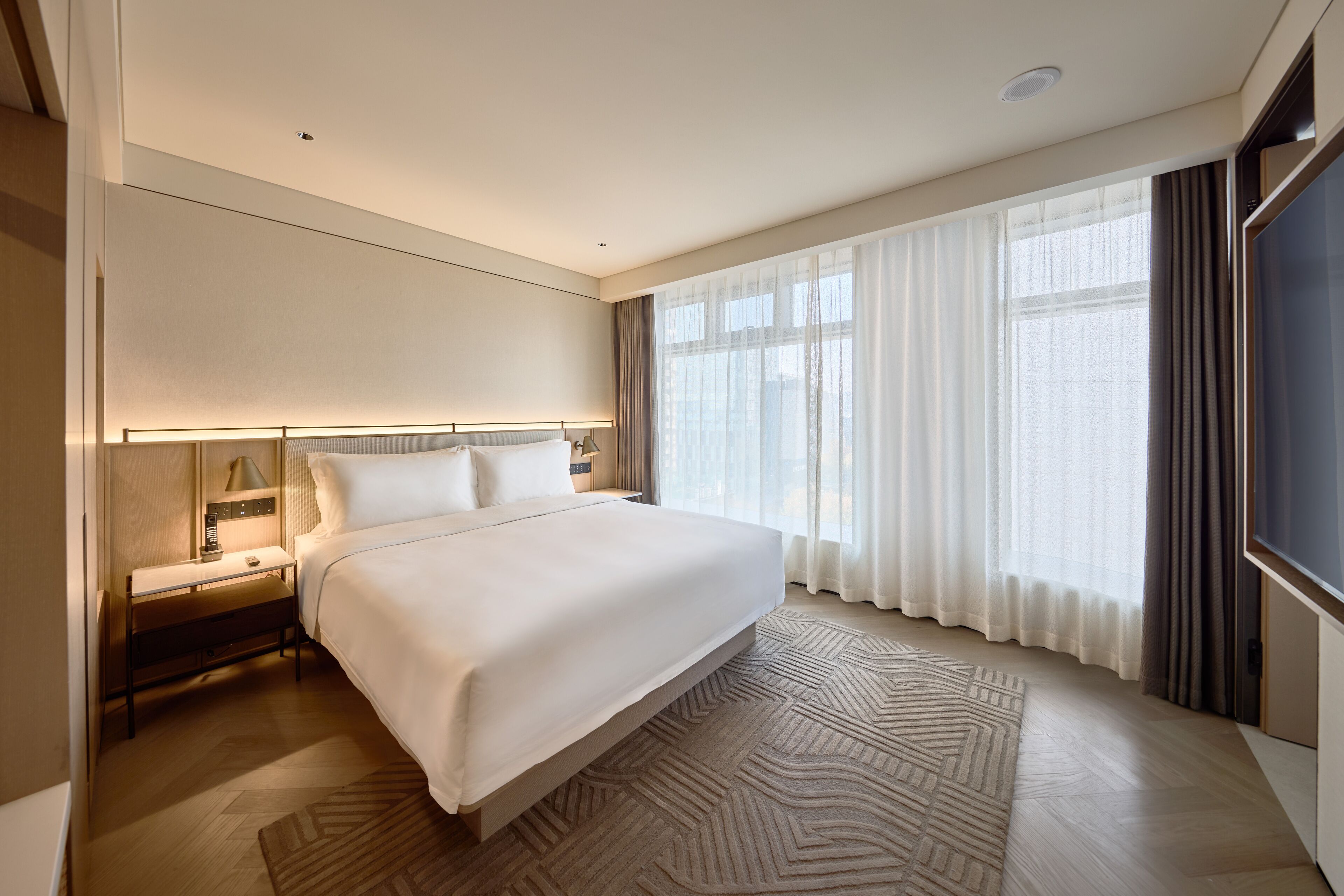 A modern hotel room with a large bed, a nightstand, a lamp, and a view of the city through large windows.